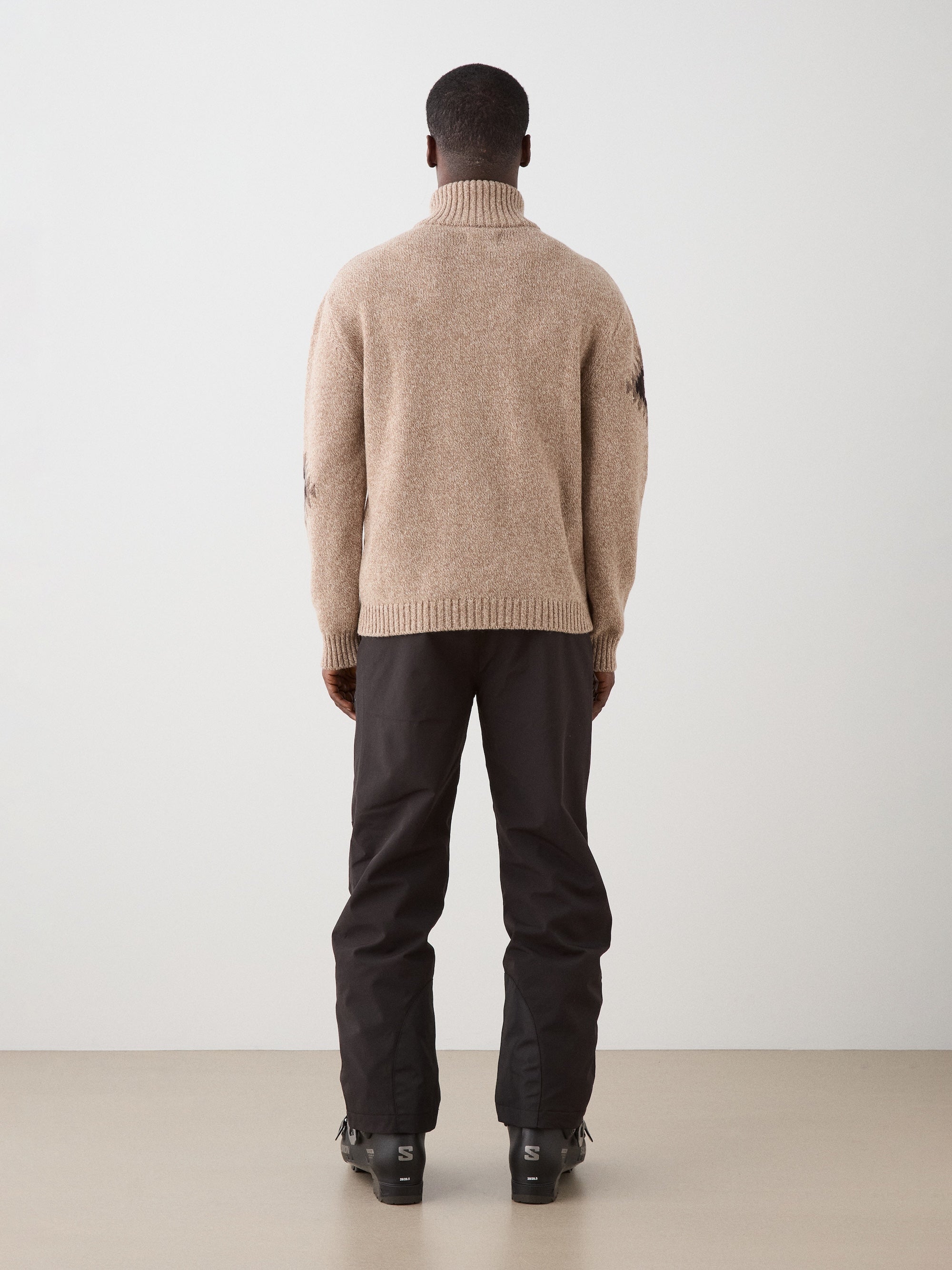 A person faces away, wearing the We Norwegians Hovden Sweater for men in beige, paired with black pants and boots against a plain, light background.