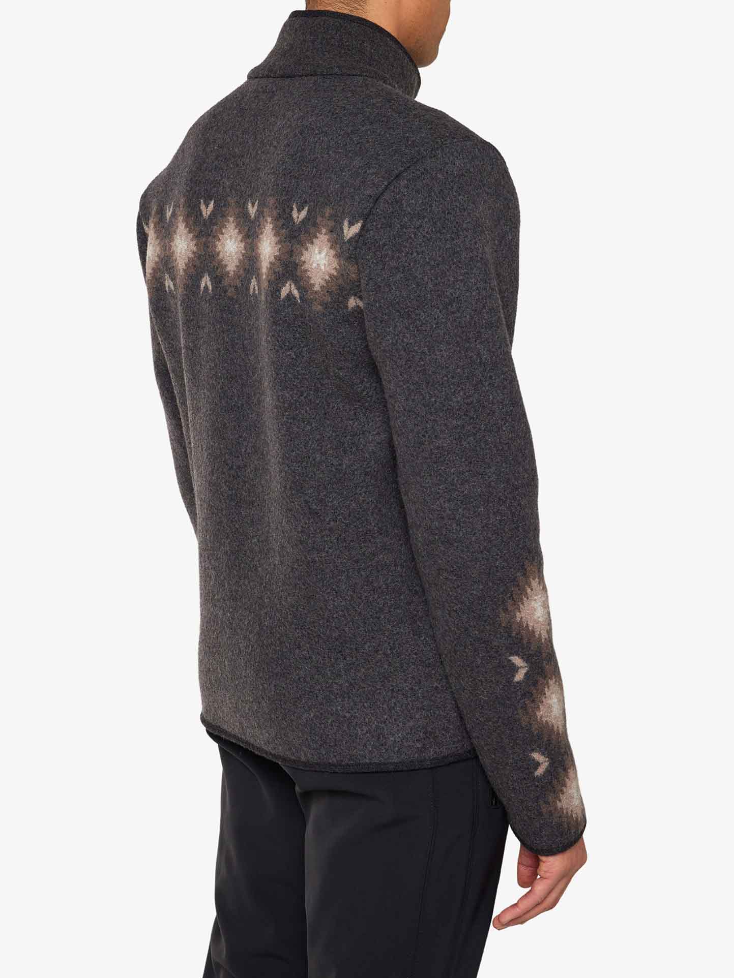 A person is shown from the back, wearing the Hovden Jacket Men by We Norwegians, a dark gray merino wool sweater with a high collar. The jacket features a pattern of light brown and white geometric designs along the upper back and near the wrists. The person is also wearing black pants.