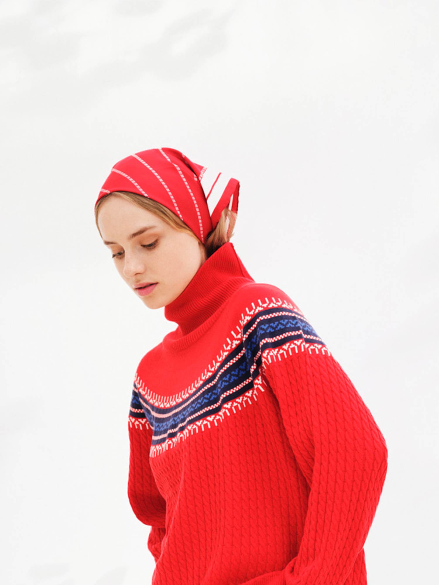 A person wearing a vibrant red sweater with intricate white and blue patterns is looking down. They have a Ship Silk Scarf Red, a red and white patterned designer accessory from We Norwegians, and the background is light and blurred, giving a soft and serene feel to the image.