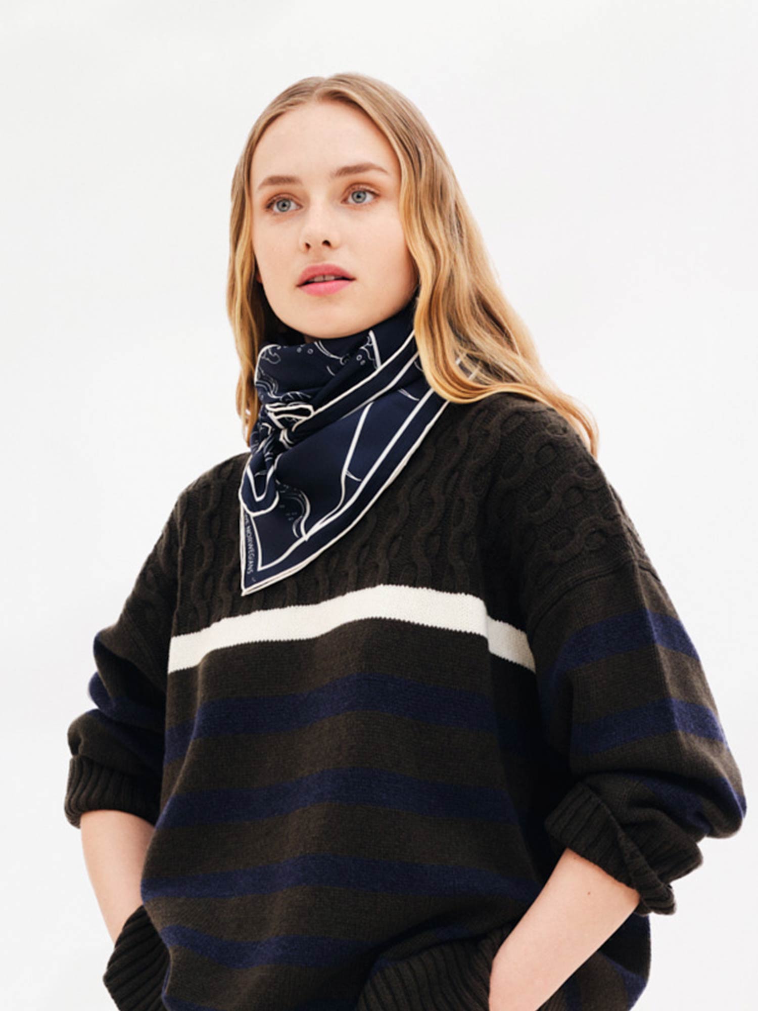 A person with long blonde hair, wearing a black, blue, and white striped sweater and a Wavy Silk Scarf Navy Blue from We Norwegians inspired by Japanese printmaking, stands against a white background. Their hands are in the pockets of their sweater, and they are gazing slightly to the side.