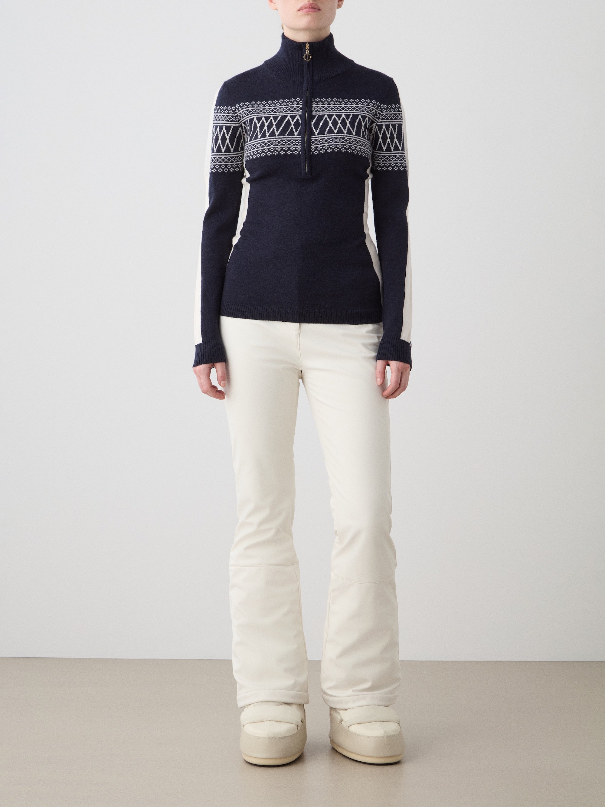 A woman stands facing forward against a plain, light background, wearing the We Norwegians Signature 1/4-Zip Women ski sweater in navy blue and white with a Setesdal pattern, off-white pants, and beige winter boots.