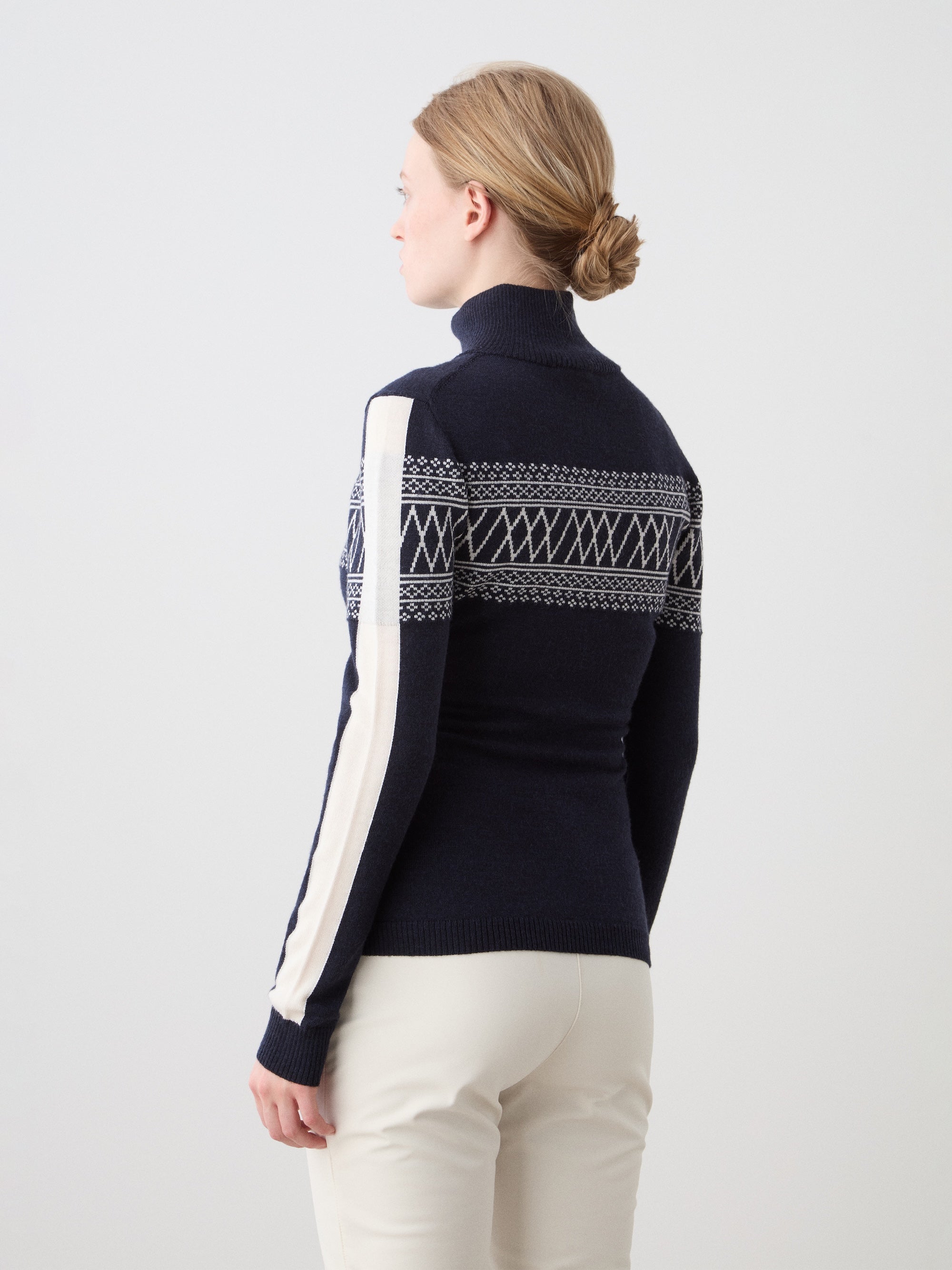 A woman with blonde hair in a low bun is seen from behind, wearing the We Norwegians Signature 1/4-Zip Women ski sweater in navy with white Setesdal-inspired patterns and striped sleeves, paired with light-colored pants.