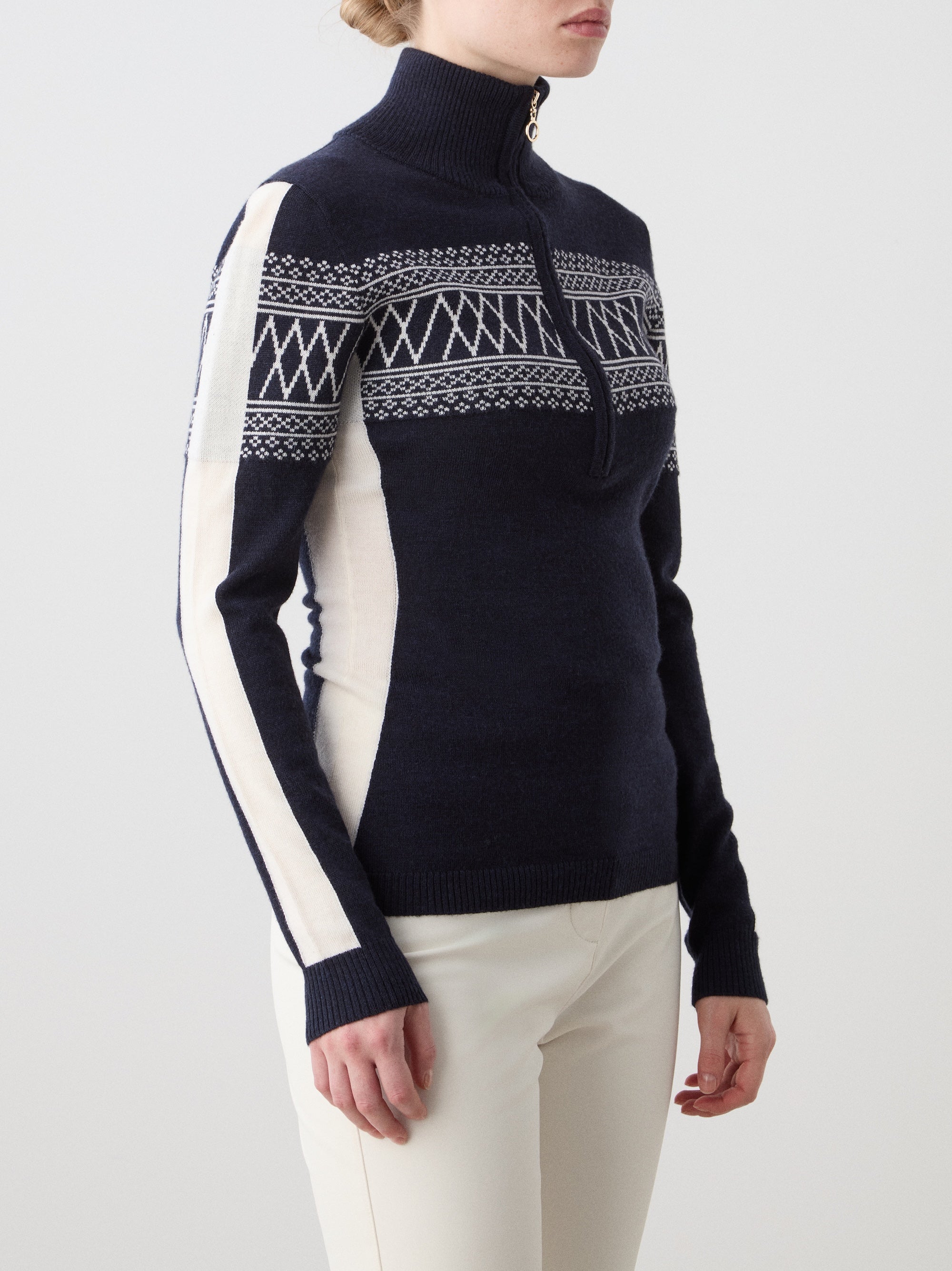 A person wears the We Norwegians Signature 1/4-Zip Women's ski sweater in navy blue with a Setesdal pattern and white sleeve stripes, paired with white pants, standing against a plain light background.