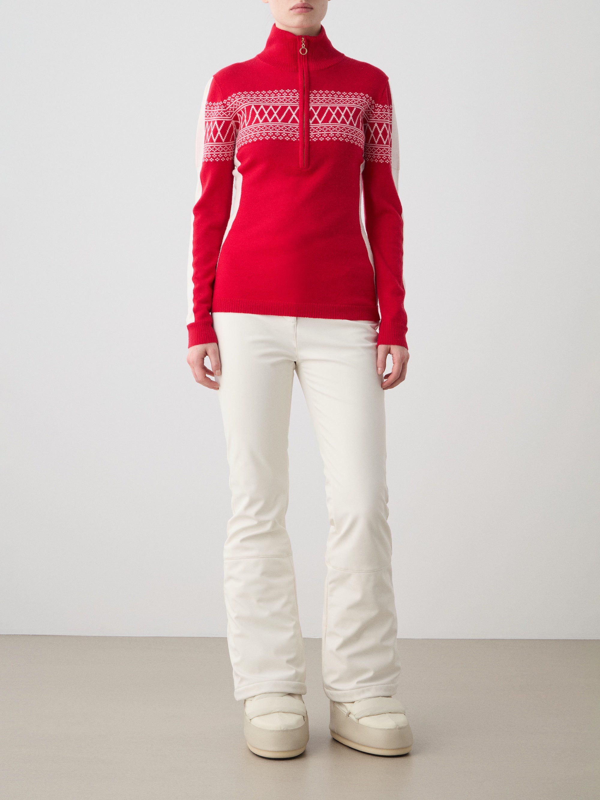 A person stands against a plain background wearing the We Norwegians Signature 1/4-Zip Women sweater with a red and white Setesdal pattern, paired with white pants and cream-colored boots. Their hands rest relaxed at their sides.