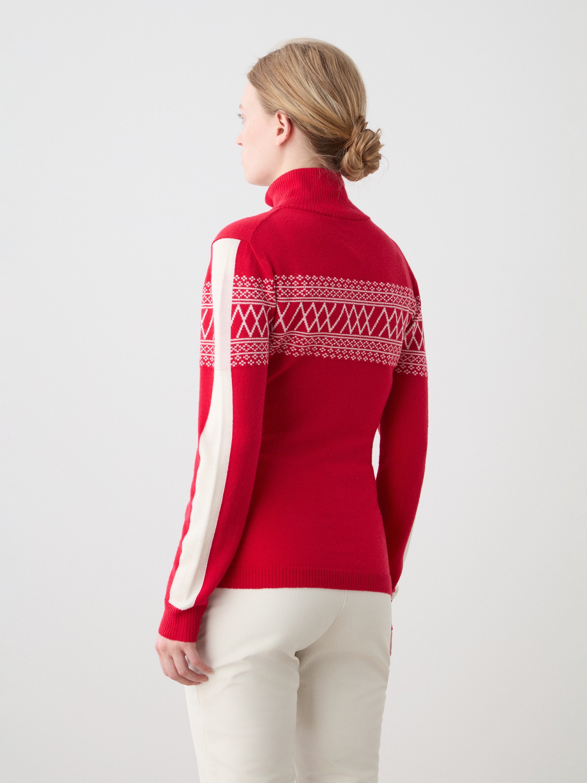 A woman with blond hair in a low bun wears the We Norwegians Signature 1/4-Zip Women ski sweater in red with white Setesdal patterns and a white sleeve stripe, paired with white pants, standing against a plain light background, facing away.