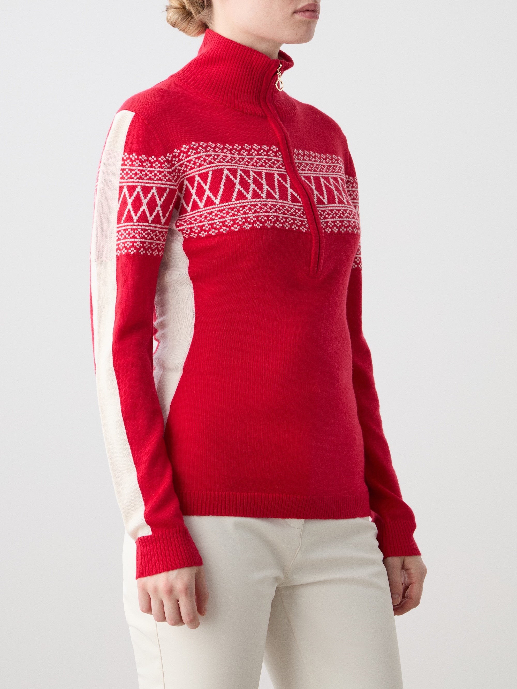 A woman wears the We Norwegians Signature 1/4-Zip Women ski sweater with a red and white Setesdal pattern and white pants, standing against a plain light background.