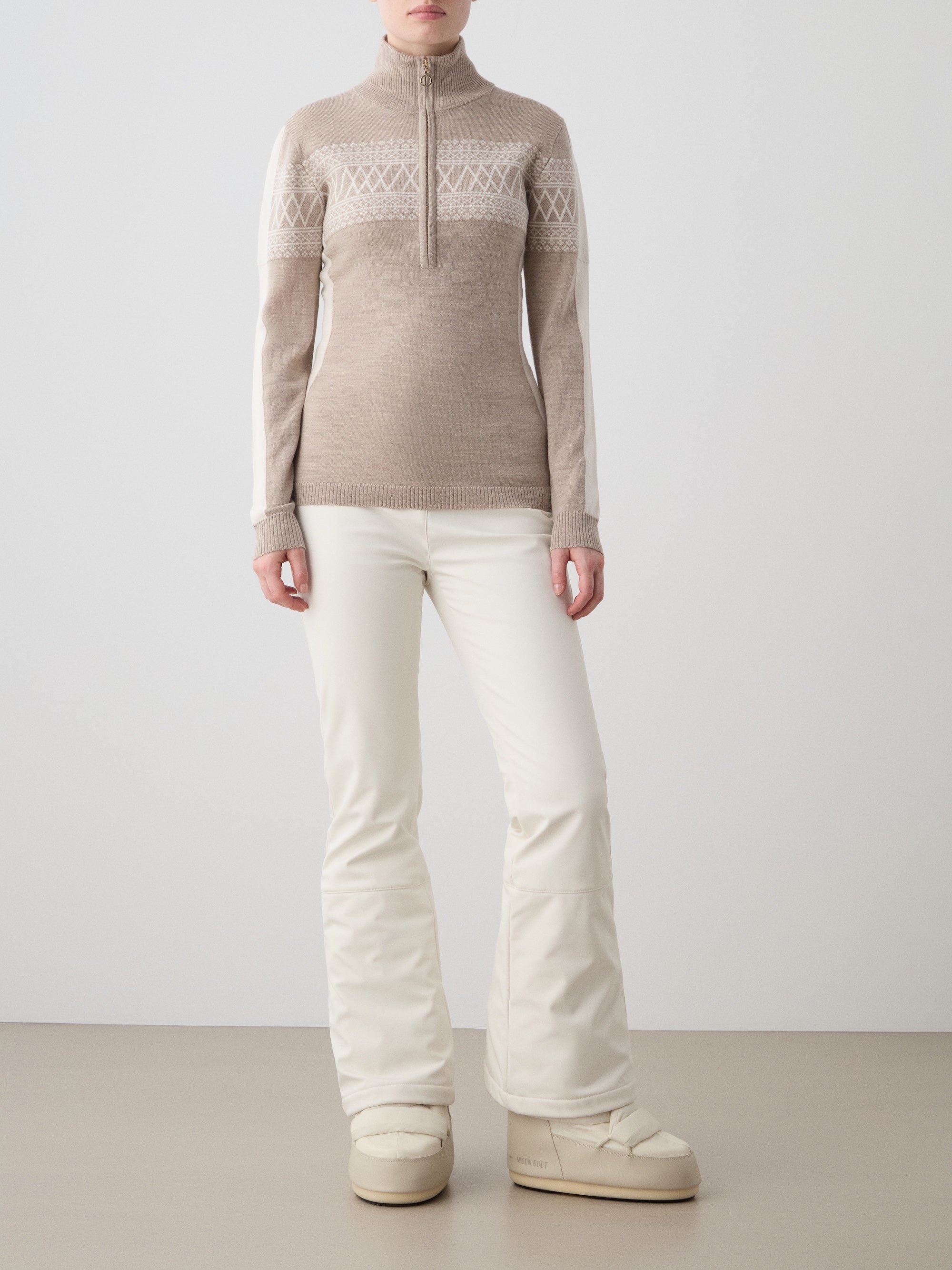 A person poses against a plain backdrop in the We Norwegians Signature 1/4-Zip Women sweater, featuring a Setesdal pattern, paired with white flared pants and beige winter boots for a chic Nordic-inspired look.