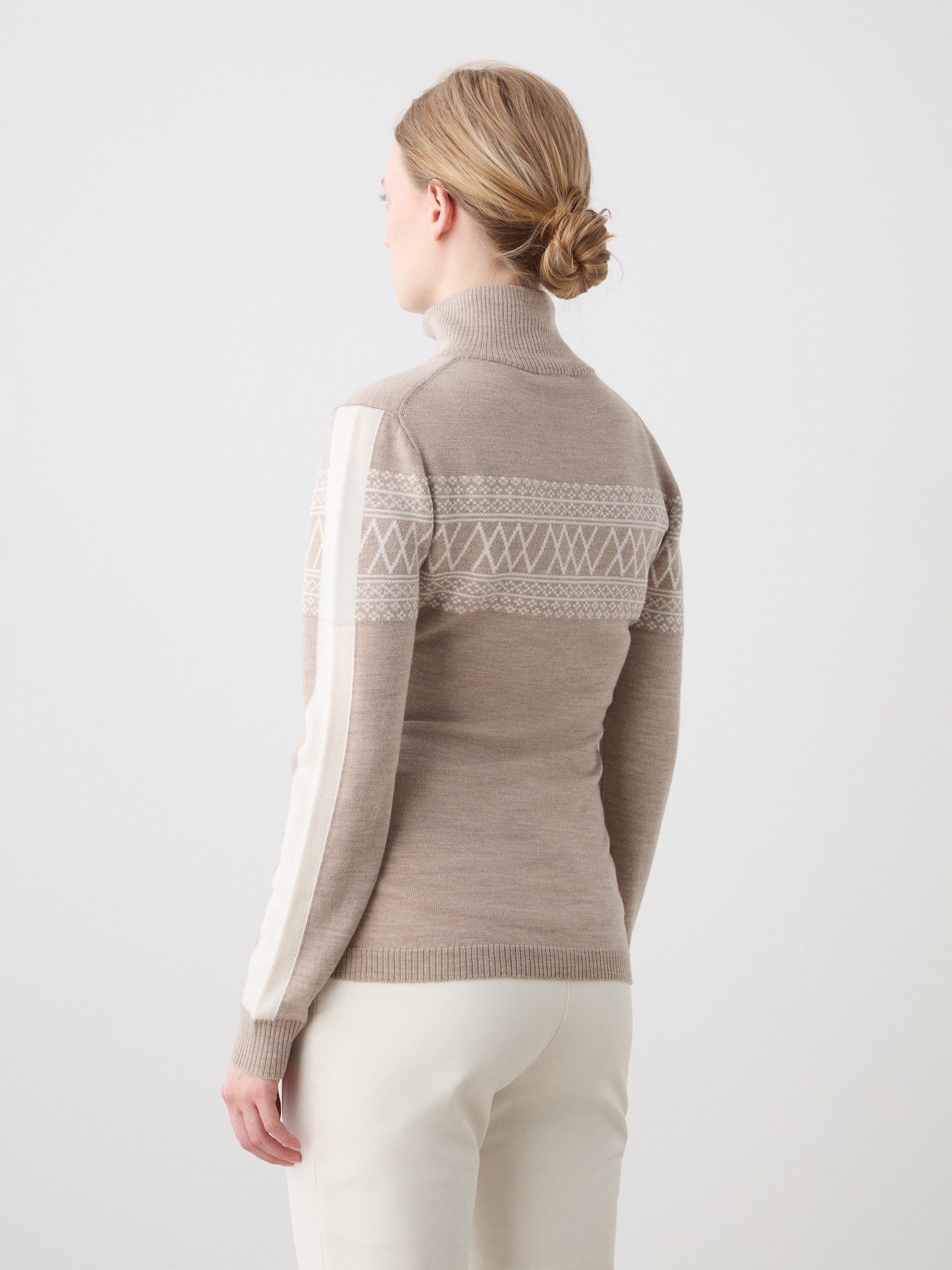 A woman with blonde hair in a low bun stands with her back to the camera, wearing the We Norwegians Signature 1/4-Zip Women ski sweater in beige with white Setesdal patterns and cream pants against a plain light background.