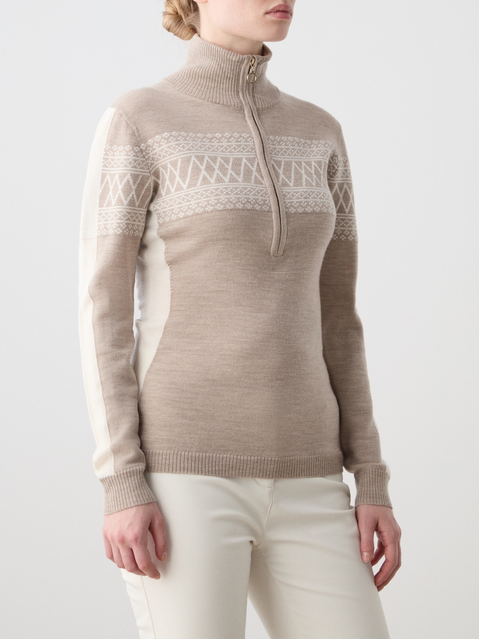 A woman wears the We Norwegians Signature 1/4-Zip Women sweater with a beige and white Setesdal pattern and half-zip turtleneck, paired with off-white pants, standing against a plain light background.