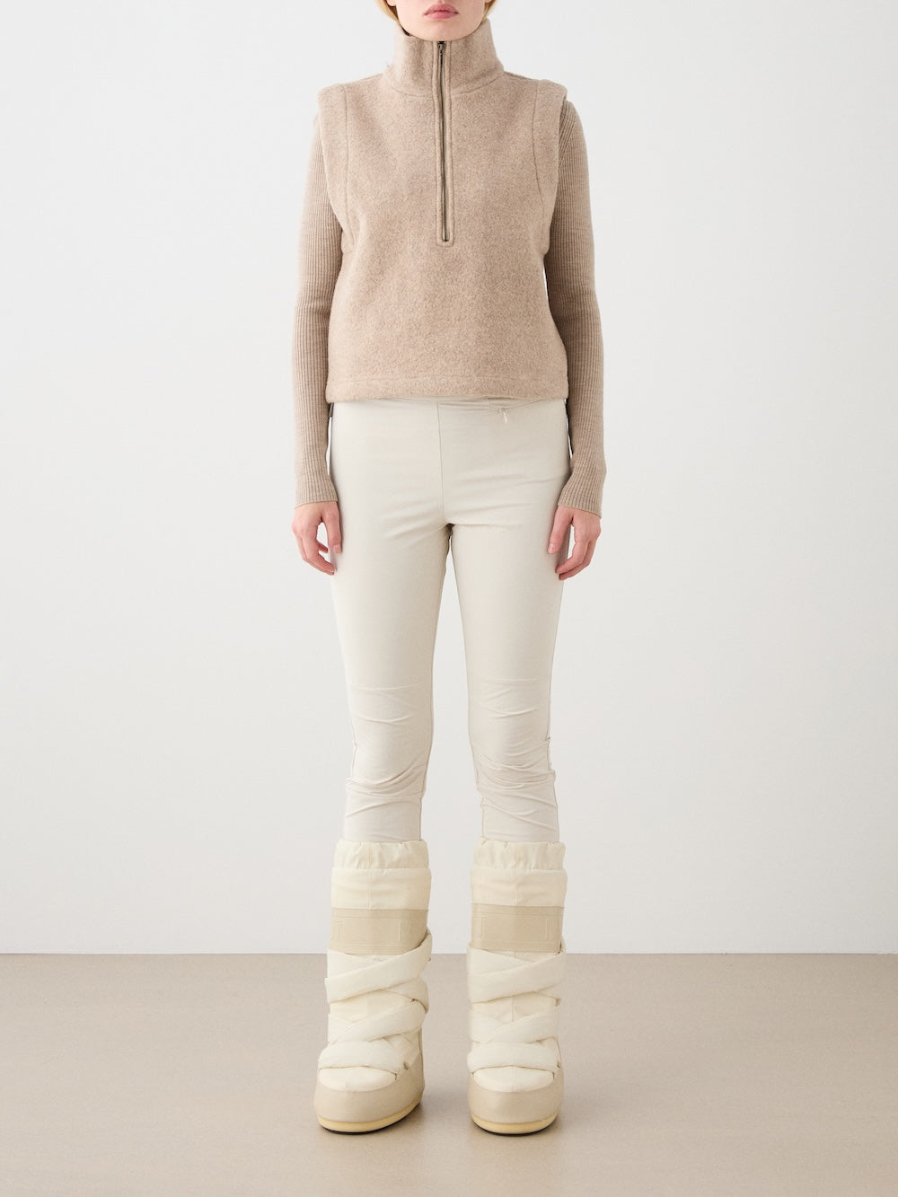 A woman stands against a plain background, styled in the We Norwegians Alta Vest Women—a beige merino wool layer worn over a light beige long-sleeve top, paired with fitted cream pants and thick off-white winter boots—embodying effortless winter style and essential layering.