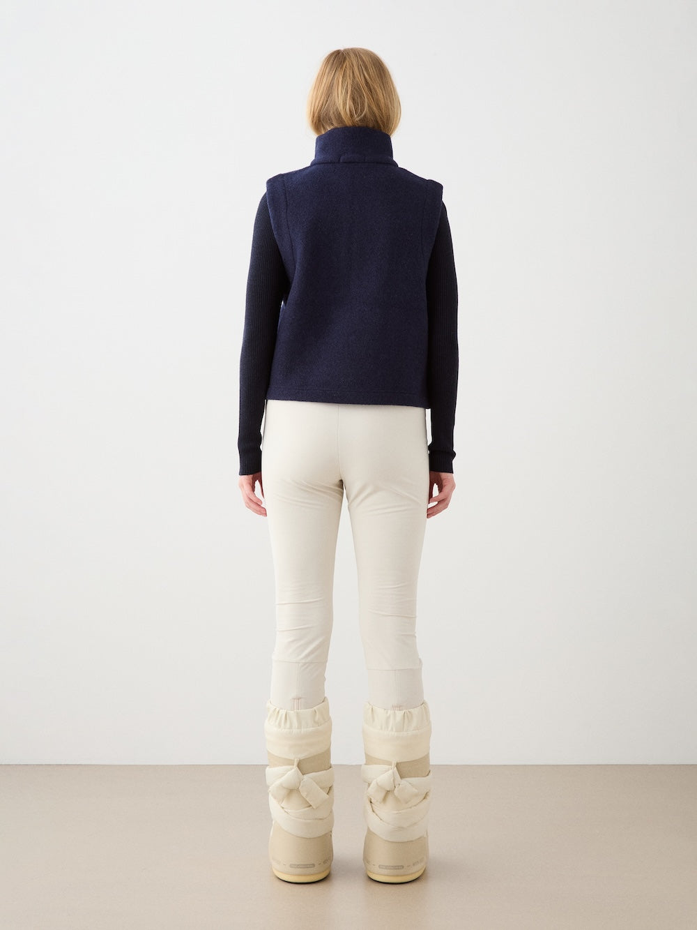A person with light brown hair, seen from behind, wears the We Norwegians Alta Vest Women in navy blue, paired with cream pants and off-white padded winter boots, set against a plain light background.
