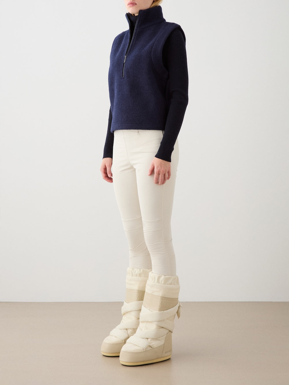 A person stands against a plain background wearing the We Norwegians Alta Vest Women—a navy merino wool layering essential—over a long-sleeve top, cream fitted pants, and tall cream puffer boots. Their head is turned slightly to the side.