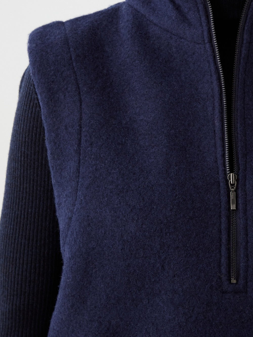 Close-up of a person wearing the We Norwegians Alta Vest Women in dark blue—a sleeveless fleece layering essential with a half-zip front—styled over a ribbed, black long-sleeve top against a plain, light background.