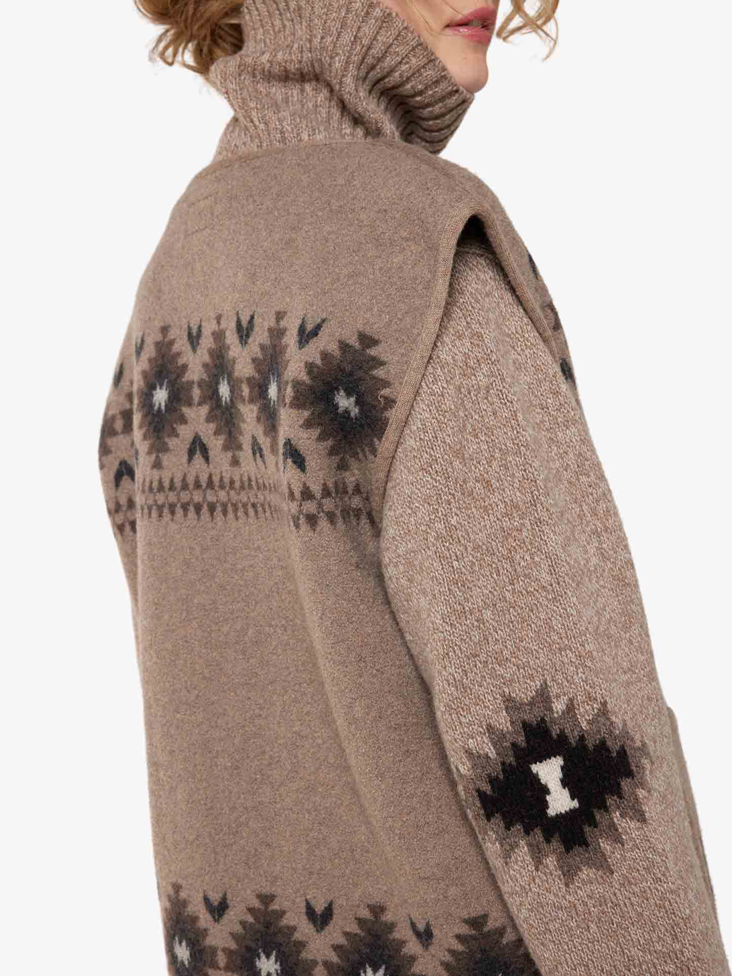 A woman is wearing the Hovden Vest Women from We Norwegians, featuring a beige turtleneck with patterned knit designs in dark and light hues on the back and sleeves. The image offers a side view from shoulder to mid-back, showcasing intricate geometric patterns inspired by Sámi Culture design.