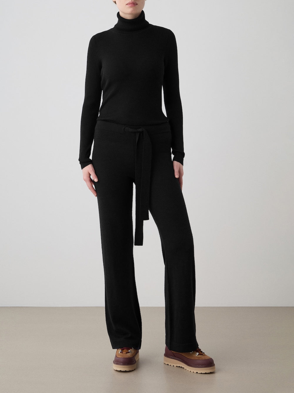 A person stands against a plain background wearing a black turtleneck sweater and We Norwegians Geilo Belted Pant Women in black, paired with brown platform shoes and hands in pockets.