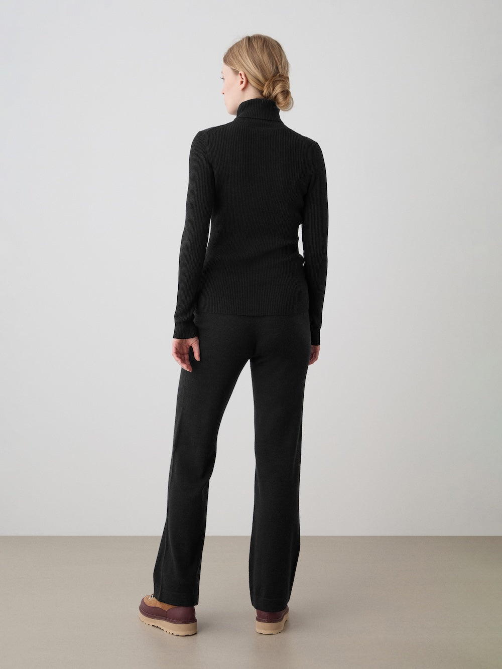 A person with blonde hair in a low bun stands facing away, wearing We Norwegians Geilo Belted Pant for women in black, paired with a black turtleneck and brown platform shoes, set against a plain light background.