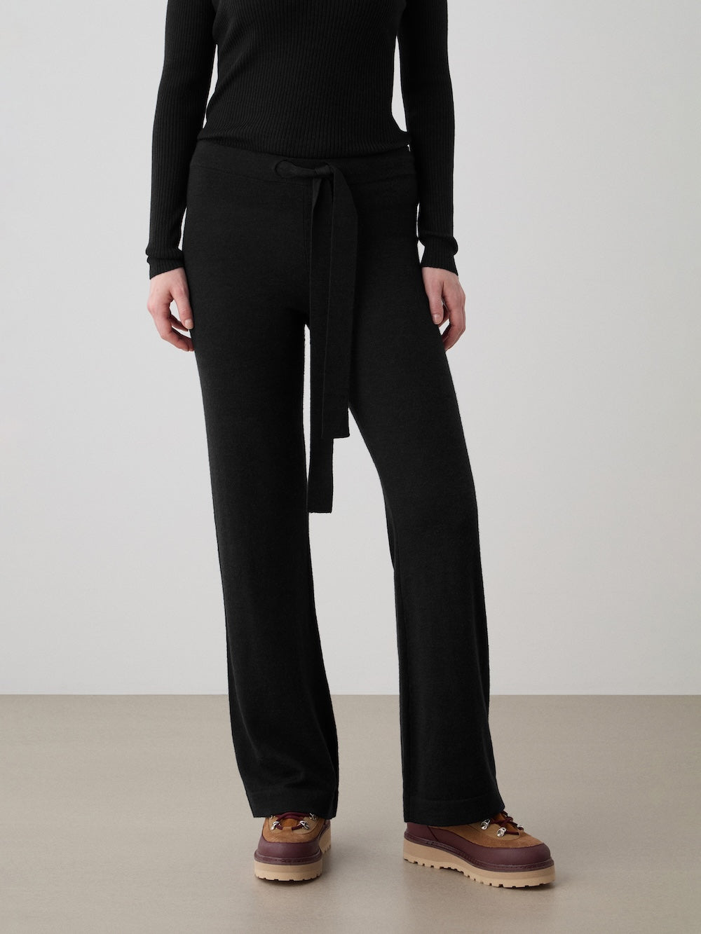 A person wears a black ribbed long-sleeve top and We Norwegians Geilo Belted Pant Women, paired with brown hiking shoes, standing against a plain background.