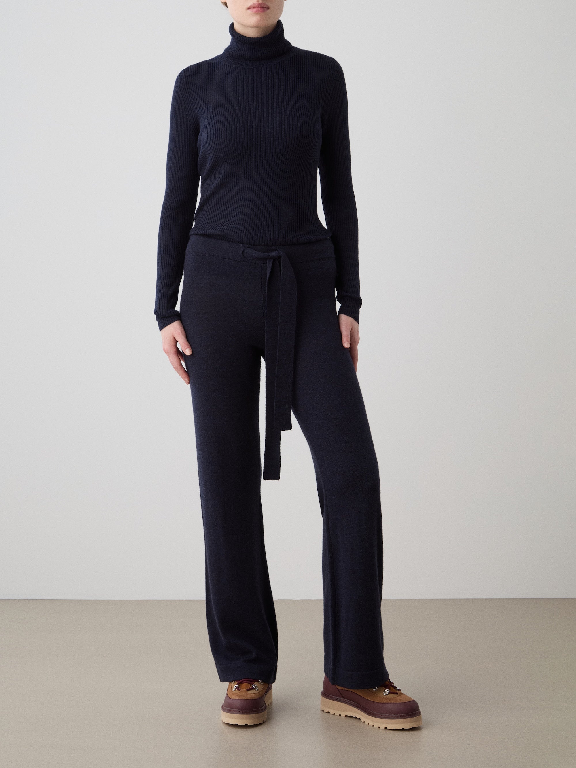 A woman wears the We Norwegians Geilo Belted Pant, styled with a dark navy turtleneck sweater and brown platform shoes, standing against a plain light gray background.