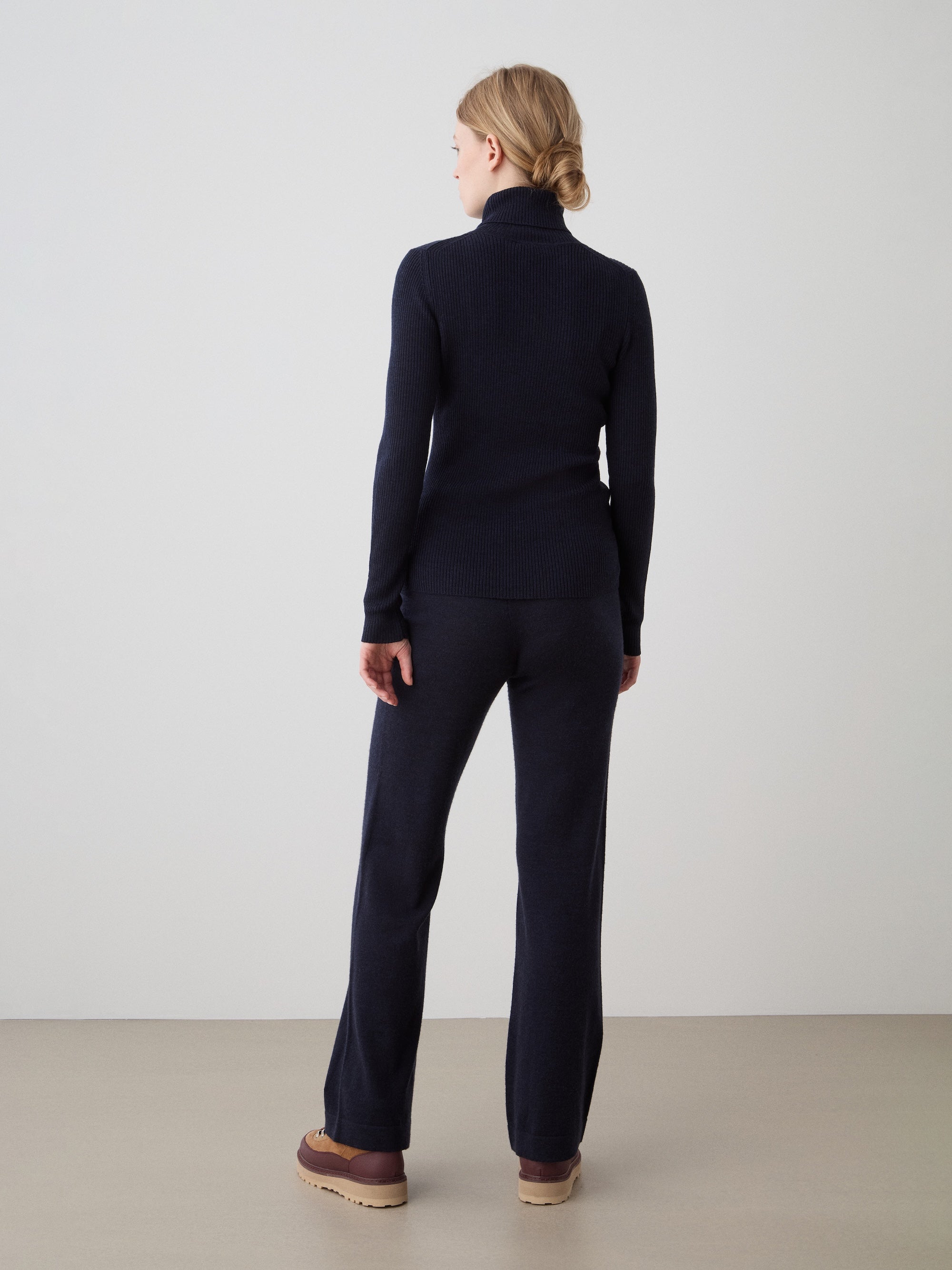 A person with blonde hair, tied back, stands facing away from the camera in a navy blue turtleneck and We Norwegians Geilo Belted Pant for women with brown shoes, set against a plain, light background.