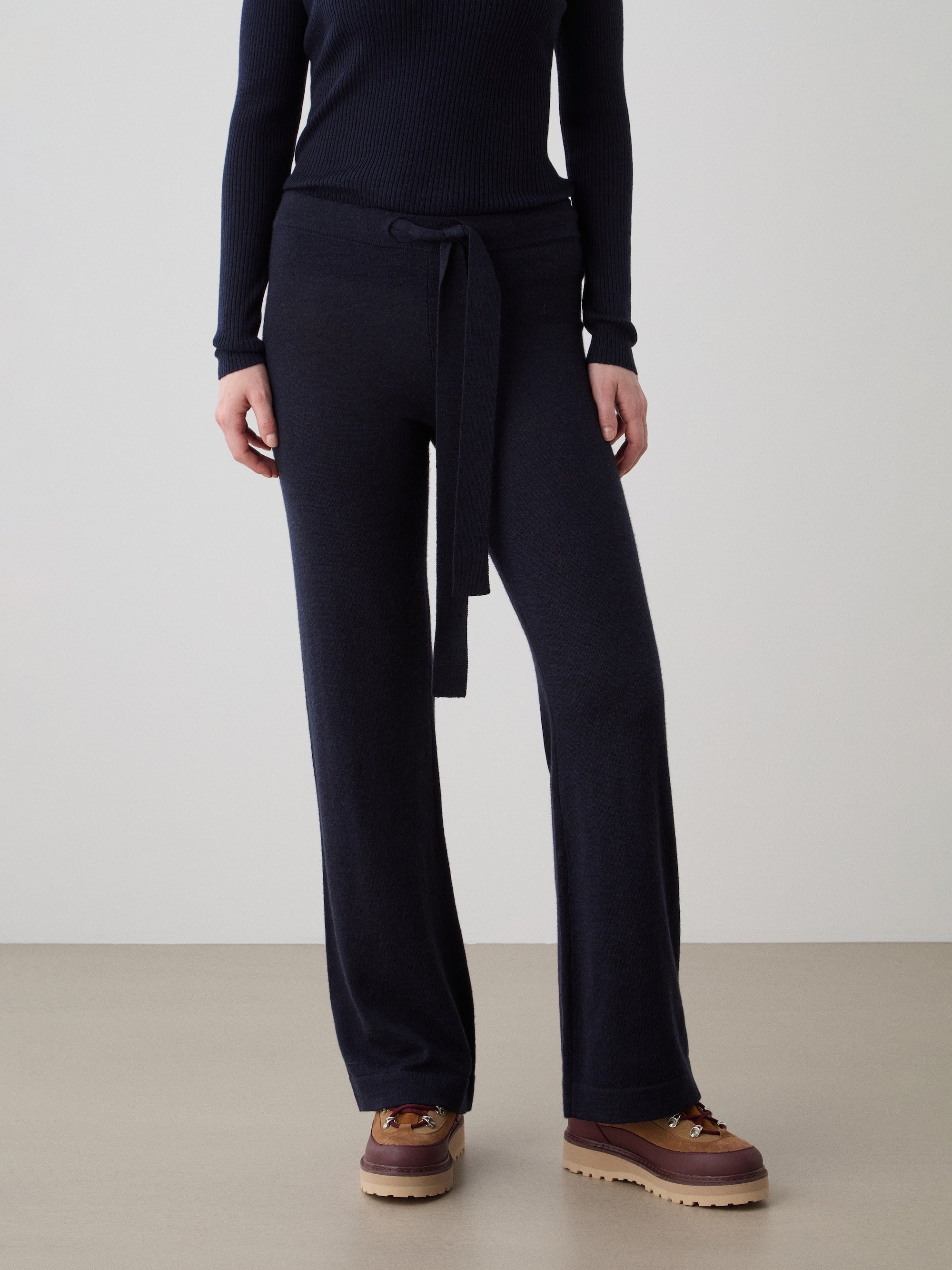 A person wears a long-sleeve black top, We Norwegians Geilo Belted Pant Women—a flared merino style with waist tie—and brown lace-up shoes, standing against a plain light backdrop.