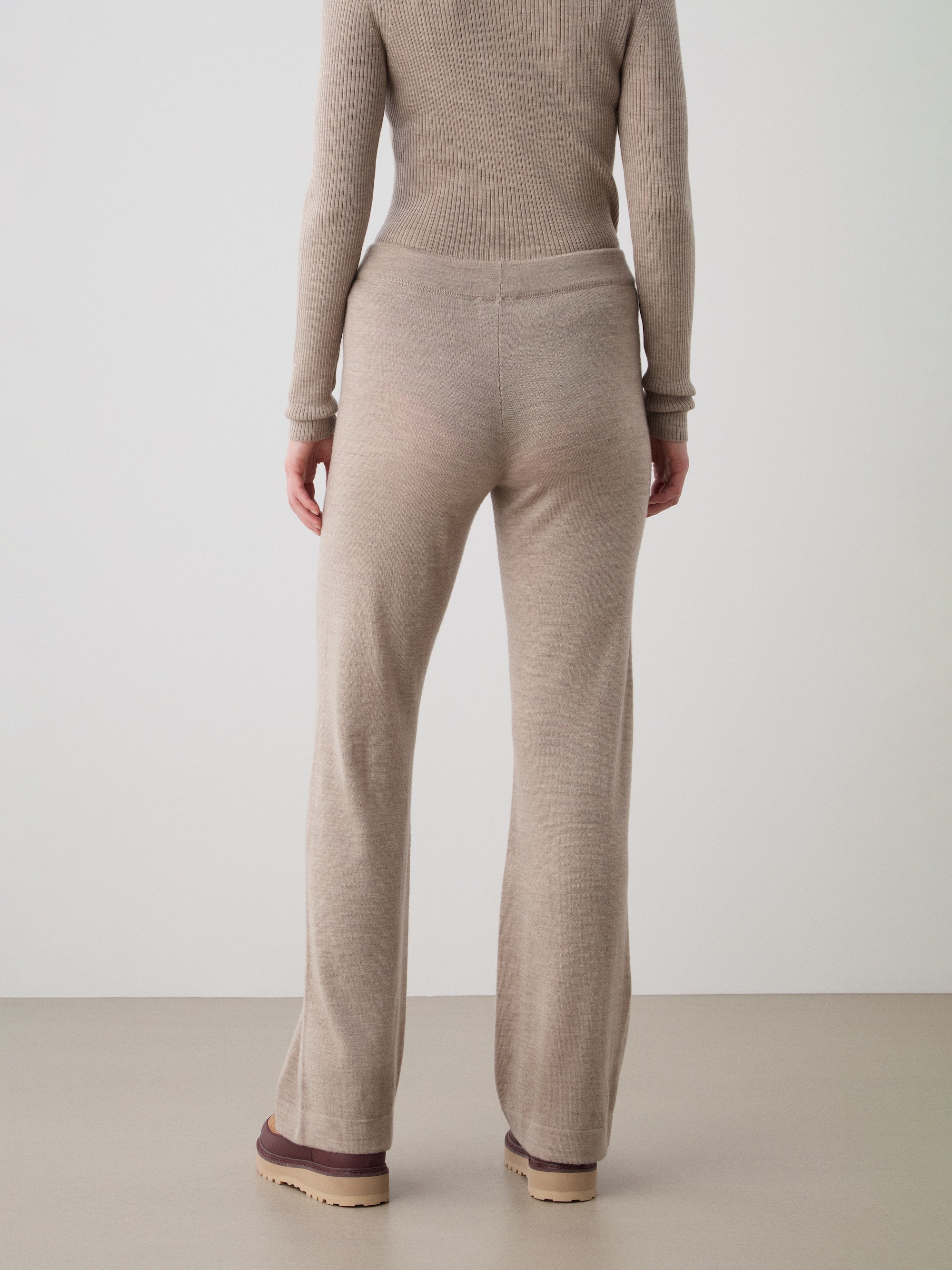 A person faces away, wearing the We Norwegians Geilo Belted Pant Women in beige with a matching ribbed top and beige shoes with thick tan soles, set against a plain light background.