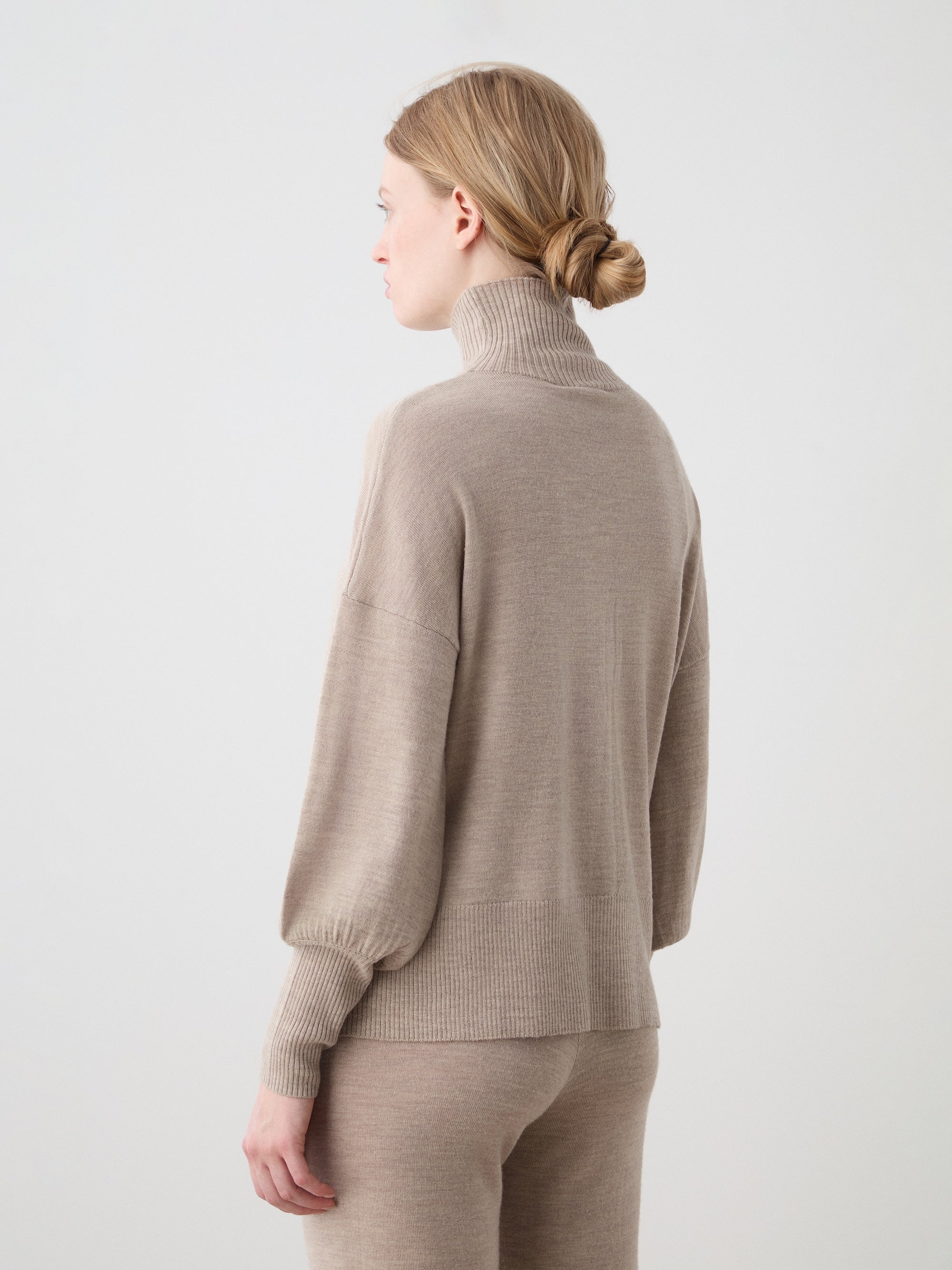 A woman with light brown hair in a low bun stands sideways, wearing the We Norwegians Geilo Oversized Sweater in beige and matching pants against a plain light background.