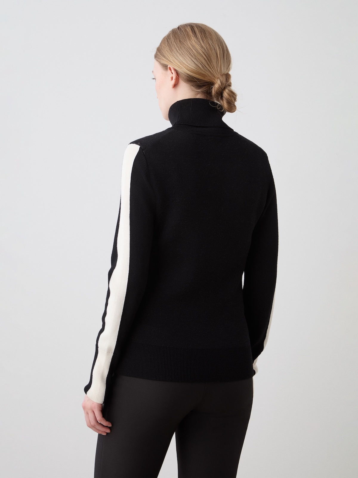 A woman with blonde hair in a low bun, seen from behind against a plain light background, wears the We Norwegians Afterski Sweater Women—black with white sleeve stripes—paired with black pants.