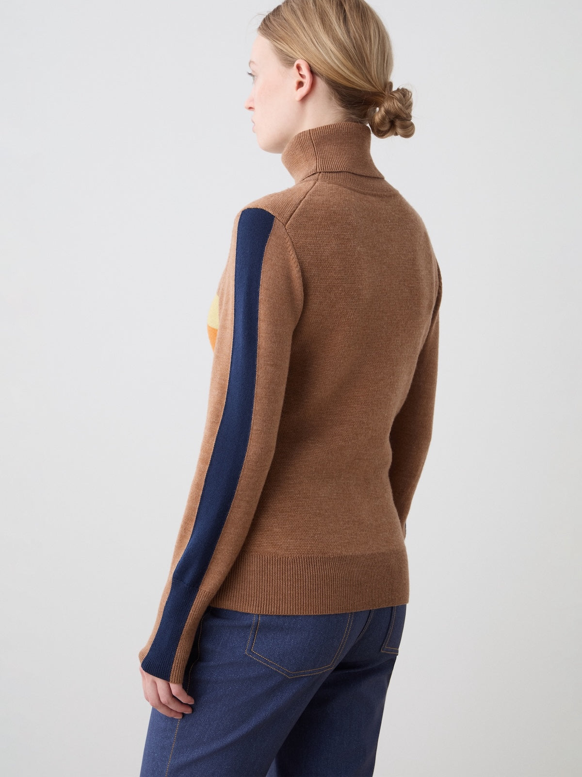 A woman with light hair in a low bun, wearing the We Norwegians Afterski Sweater Women (brown with a navy sleeve stripe) and blue jeans, stands facing away from the camera against a plain light background.