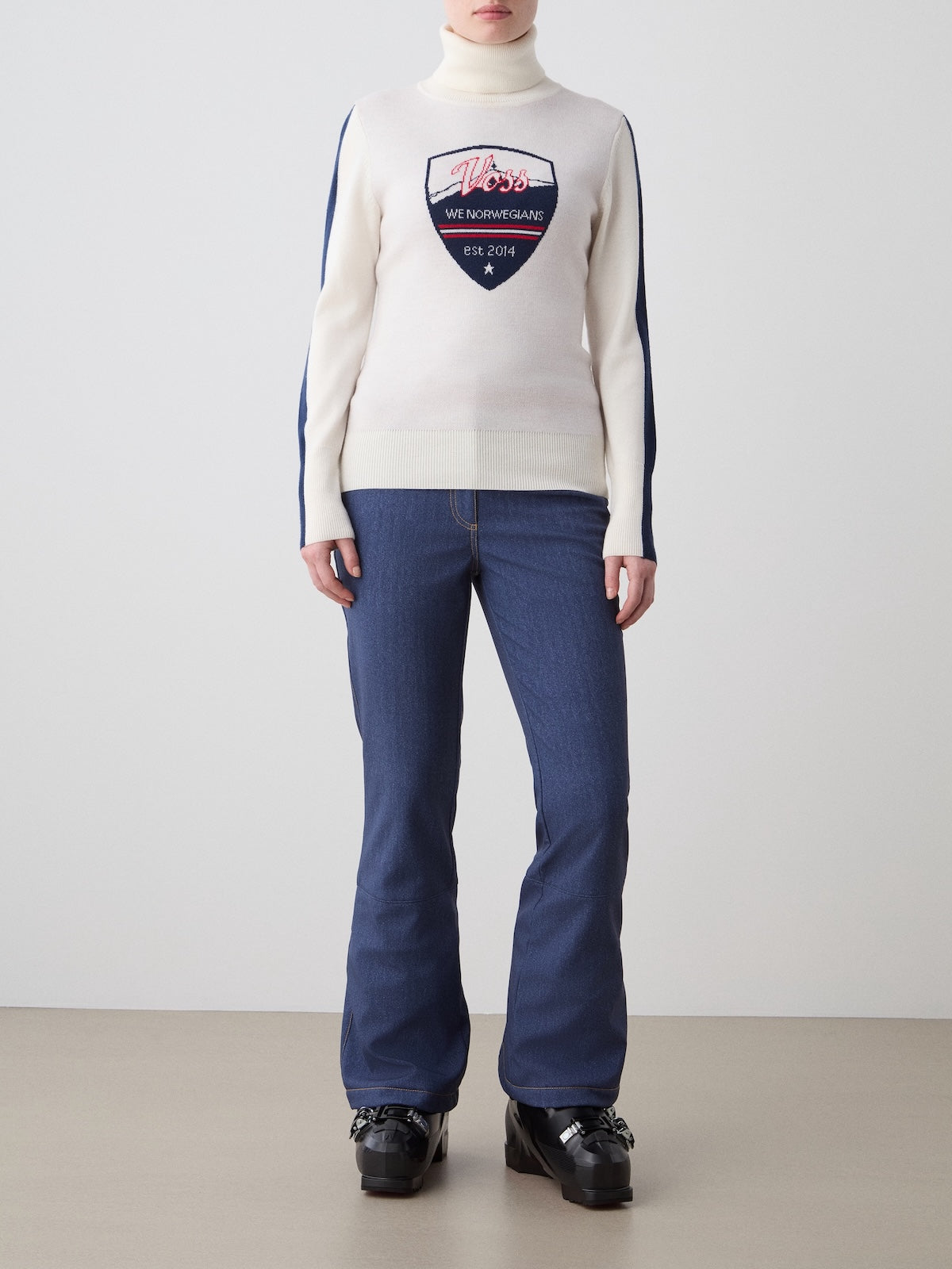 A person stands indoors against a plain background wearing the We Norwegians Afterski Sweater Women—a white retro-style top with a navy emblem and text—paired with blue flared jeans and black platform shoes.