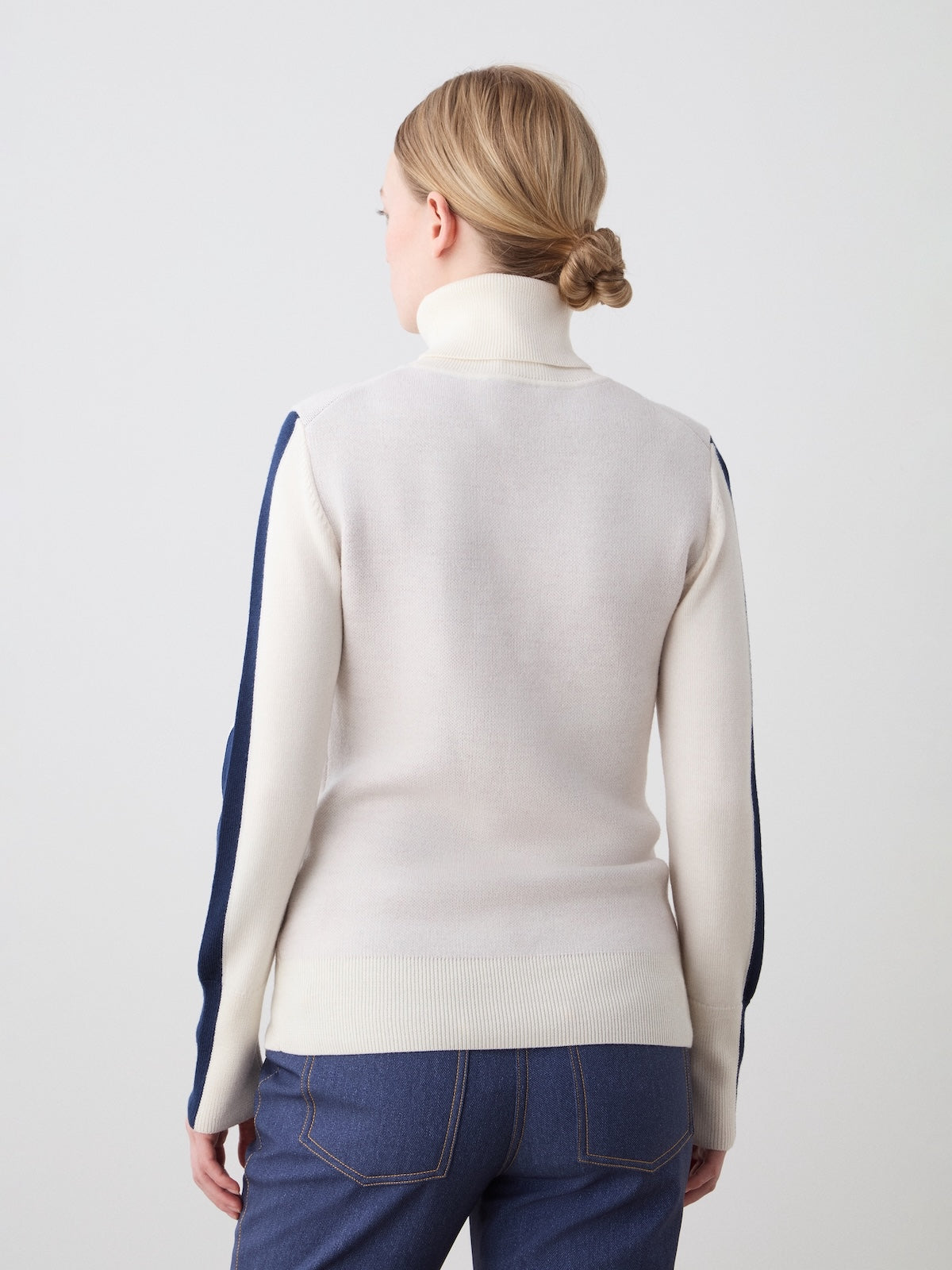 A woman with blonde hair in a low bun is seen from the back, wearing the We Norwegians Afterski Sweater Women in cream with navy sleeve stripes and blue jeans, standing against a plain light background.