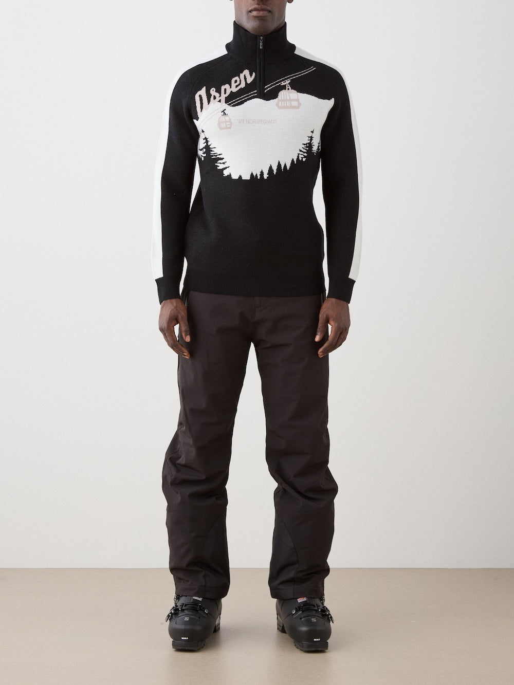 A person wearing the We Norwegians Afterski Sweater Men, featuring a mountain and cable car design, along with black ski pants and black ski boots, stands against a plain light-colored wall.