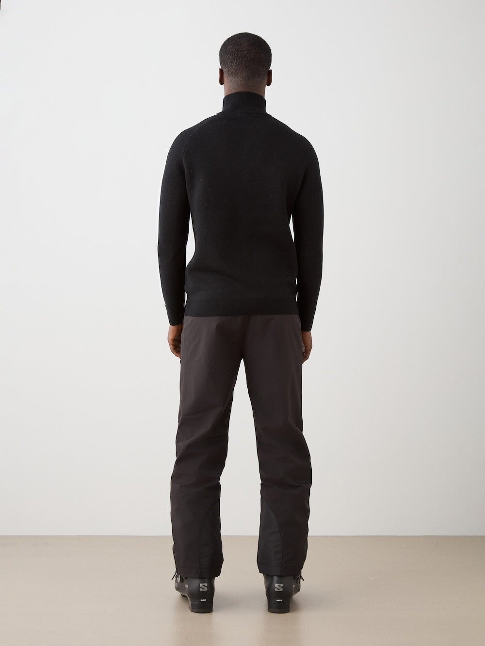 A man faces away from the camera, wearing the We Norwegians Afterski Sweater Men in black with dark pants, set against a plain light background.