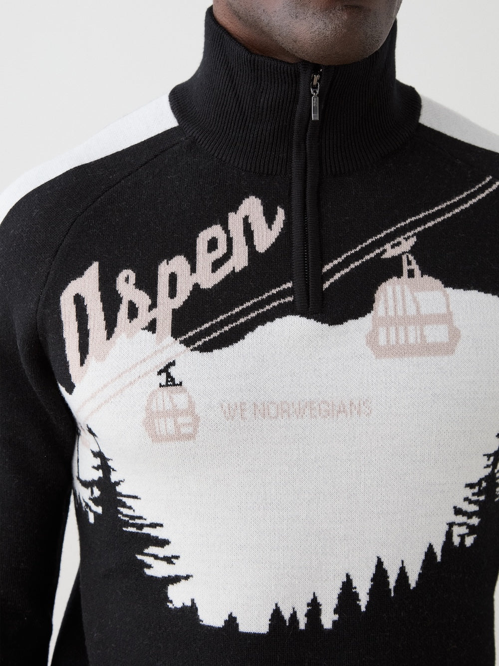 A close-up of a person wearing the We Norwegians Afterski Sweater Men, a black and white merino wool ski sweater with "Aspen," ski gondolas, pine trees, and "WE NORWEGIANS" knit into the high collar design with a short front zipper.