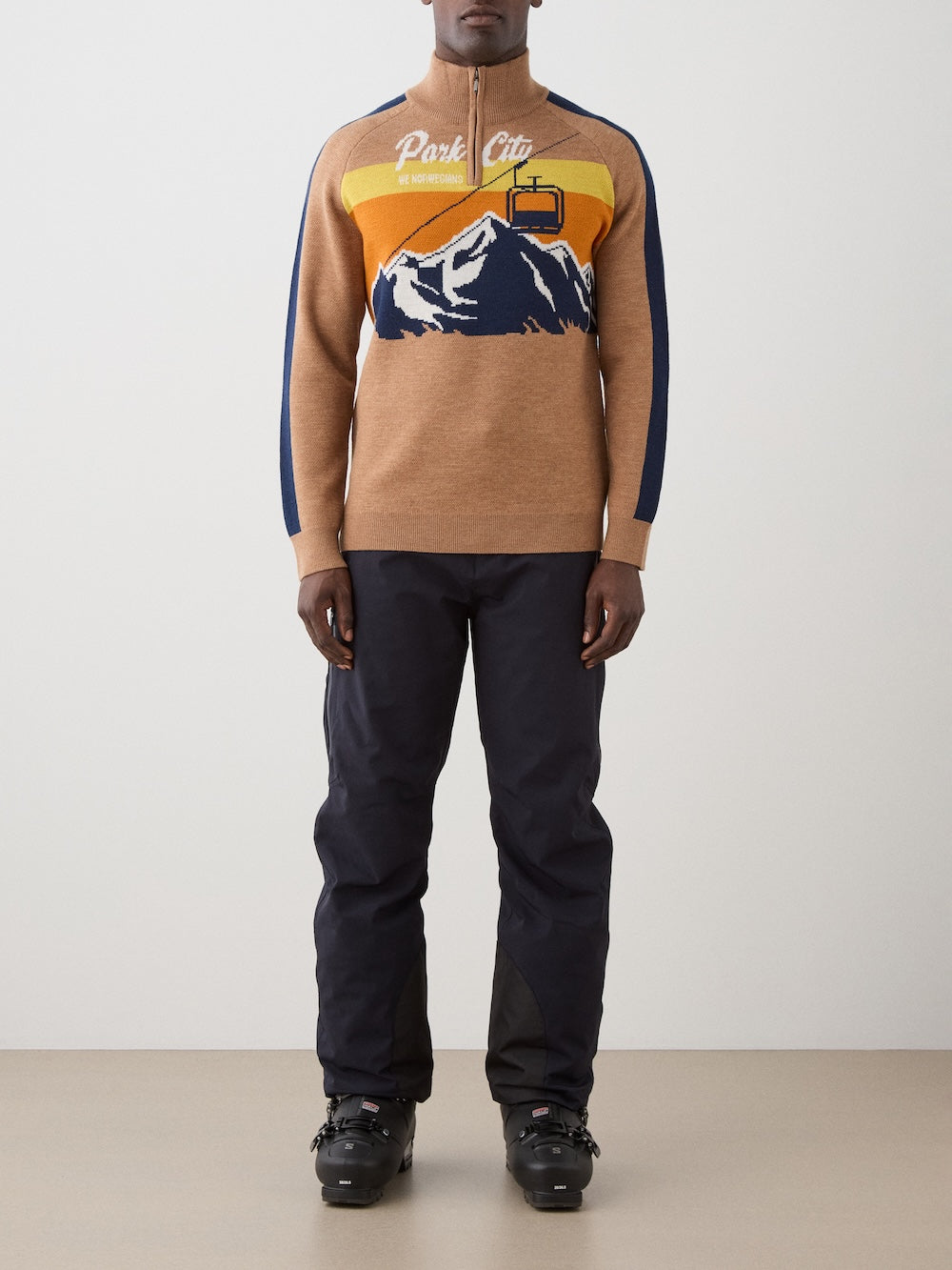 A man wears the We Norwegians Afterski Sweater Men featuring a mountain, cable car, and "Park City" graphic, paired with black pants and ski boots, standing against a plain light background.