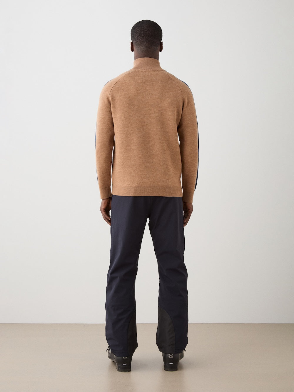 A man faces away from the camera, wearing the We Norwegians Afterski Sweater Men in brown, paired with dark pants and black shoes, set against a plain white wall and beige floor.