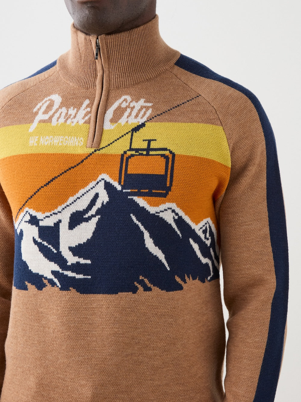A man wears the We Norwegians Afterski Sweater Men in brown, featuring snowy mountains, a ski lift, "Park City" text, and navy and yellow stripes—a classic vintage look.