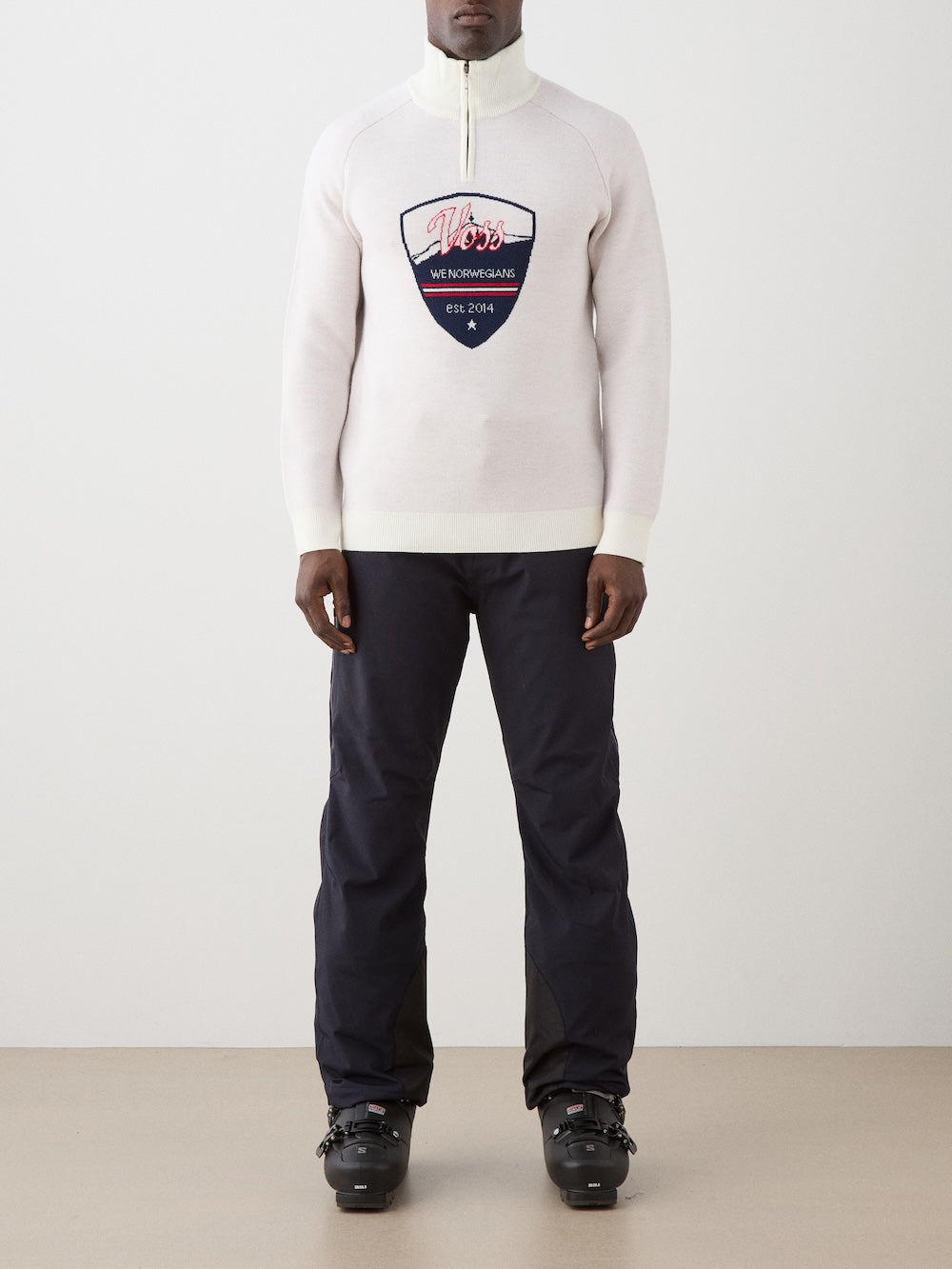 A person indoors wears the We Norwegians Afterski Sweater Men—white merino wool with a large blue and red chest patch—paired with black pants and black ski boots. The background is plain and light-colored.