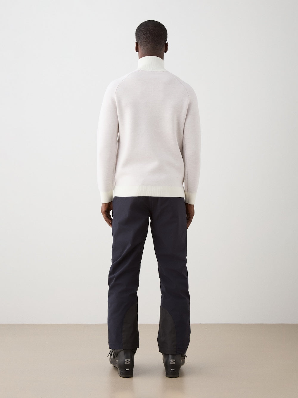 A man stands facing a white wall, seen from behind, wearing the We Norwegians Afterski Sweater Men with dark pants and black boots on a light-colored floor.