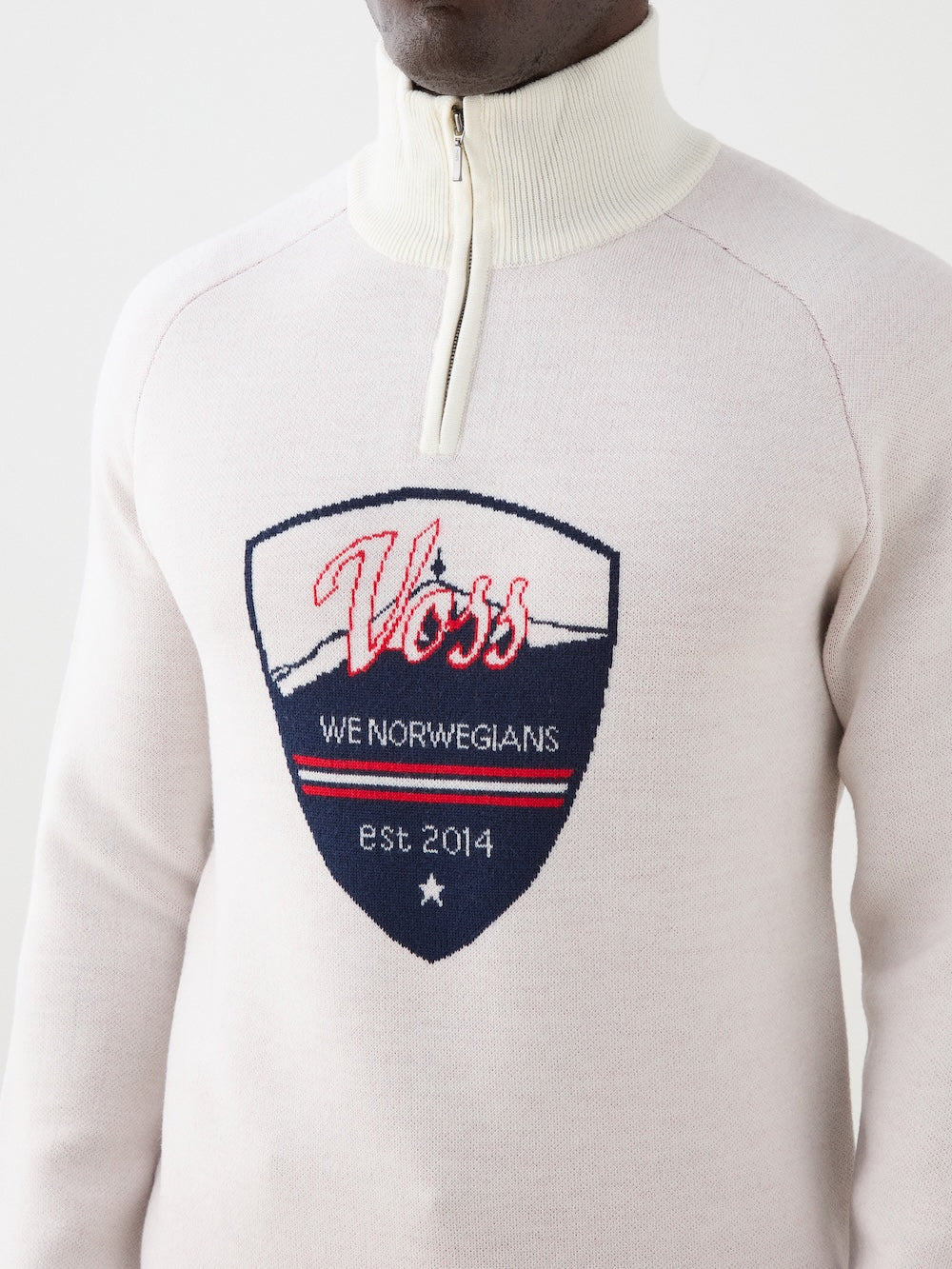 A person wearing the We Norwegians Afterski Sweater Men, a light-colored vintage zip-neck with a large "Voss We Norwegians est 2014" embroidered patch in red, white, and blue, featuring a star and mountain design.