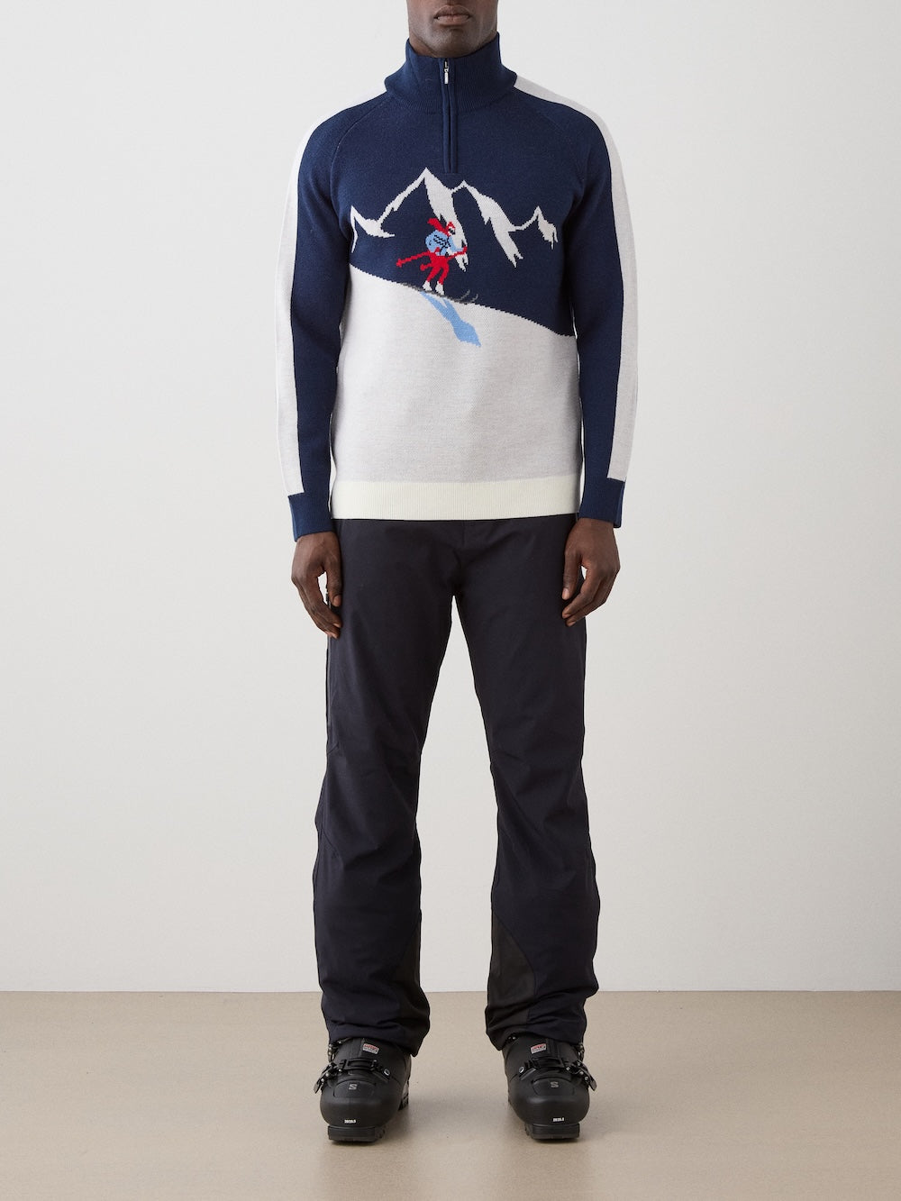 A person wearing the We Norwegians Afterski Sweater Men—a navy and white retro ski sweater featuring a mountain and skier design—paired with black ski pants and black ski boots stands against a plain background.