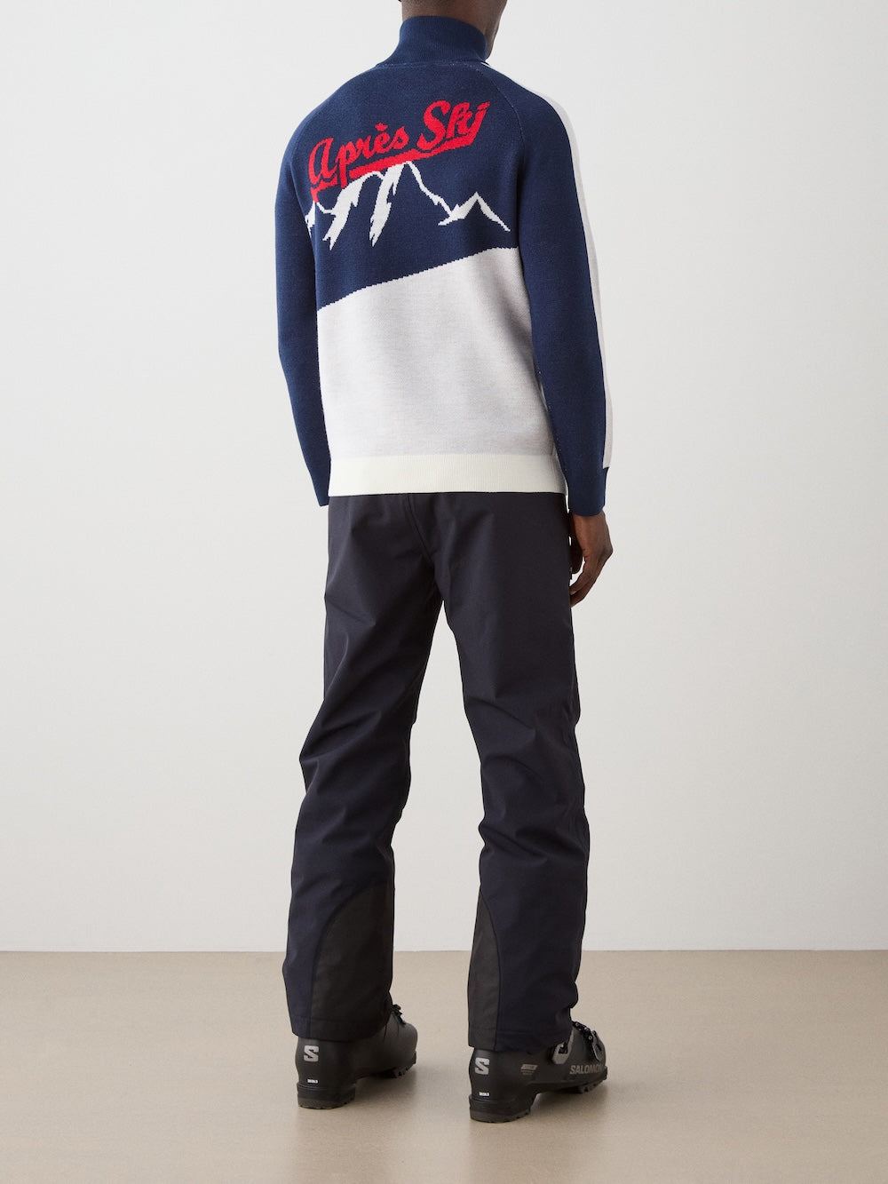 A person stands facing away, wearing the We Norwegians Afterski Sweater Men featuring a mountain graphic and red "Après Ski" text on the back, paired with dark pants and black ski boots, on a beige floor against a plain wall.