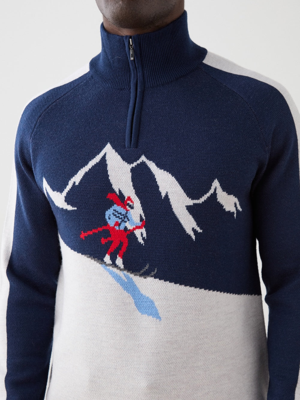 A person wears the We Norwegians Afterski Sweater Men, a navy and white zip-neck knit with a red and blue skier on a snowy mountain design.