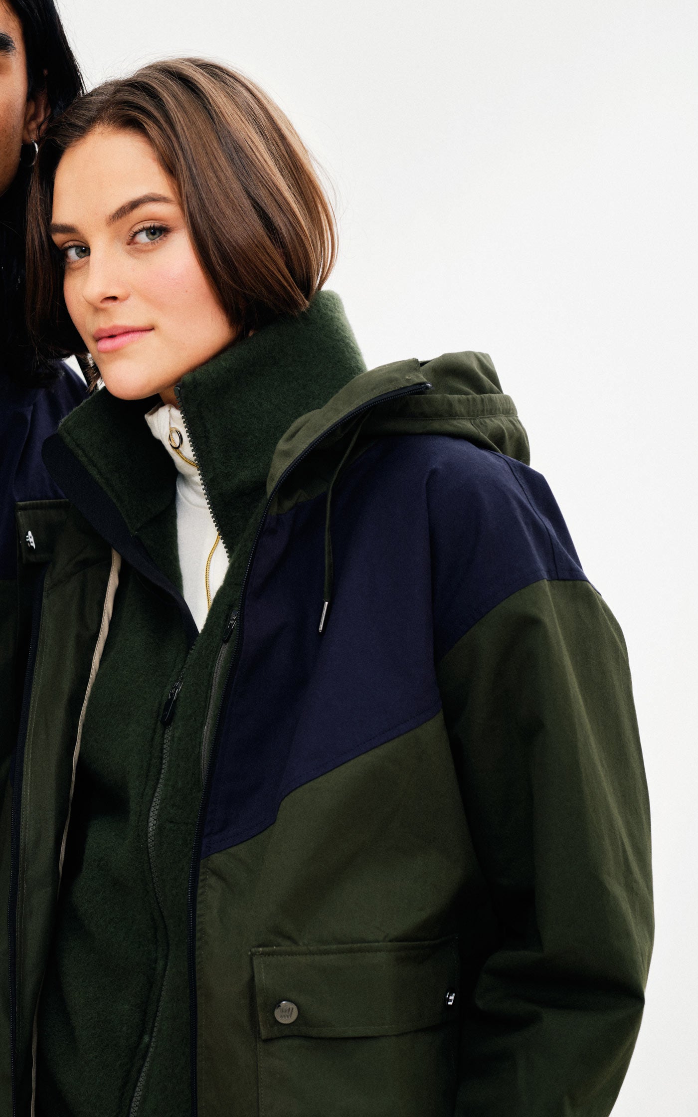 A person with short brown hair is wearing the Lyngen Jacket Women Olive Green by We Norwegians, made from Ventile® cotton fabric. They have a slight smile and are looking into the camera. The background is plain and white, emphasizing the outdoor enthusiast's face and weatherproof attire.