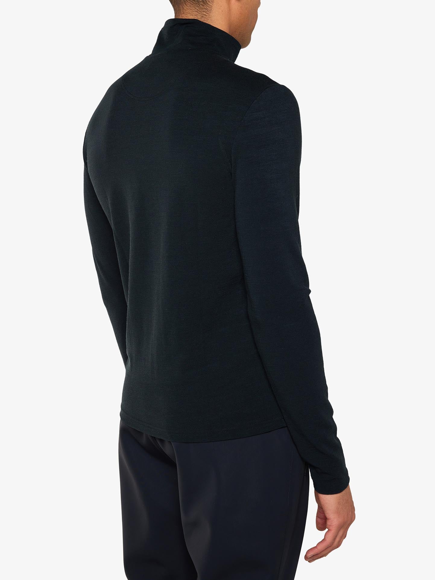 The image features a person from behind, clad in a long-sleeve, dark-colored turtleneck sweater and dark pants. The sweater, identified as the Voss Chevron Zipup Men from We Norwegians, is crafted from Superfine Merino wool and radiates retro vibes. The plain white background accentuates the relaxed fit and premium quality of this elegant garment.