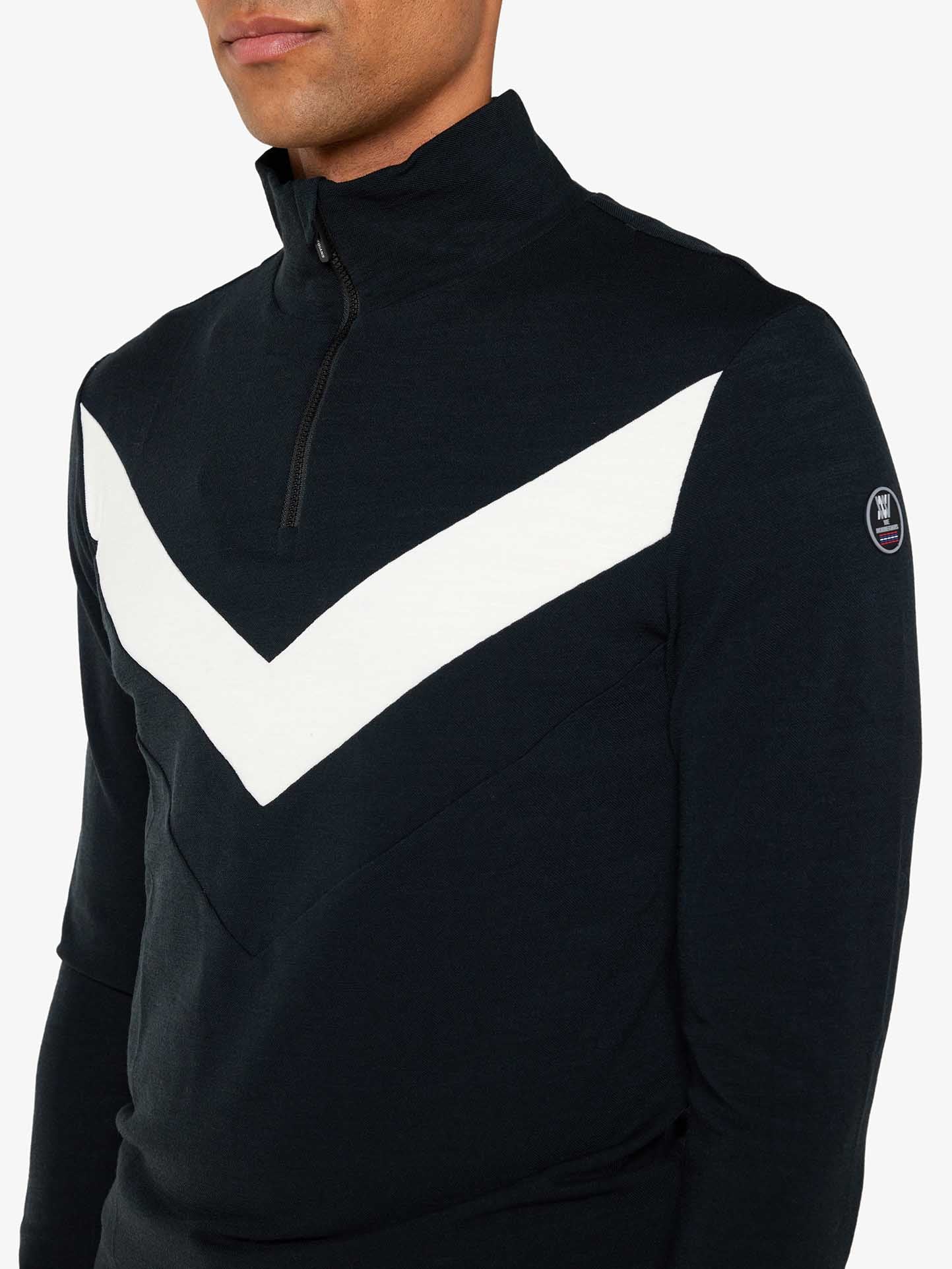 A person wears a black We Norwegians Voss Chevron Zipup Men pullover, featuring a white V-shaped design on the chest that exudes retro vibes. The pullover has a high collar and a small logo patch on the left sleeve, while the background is plain white.