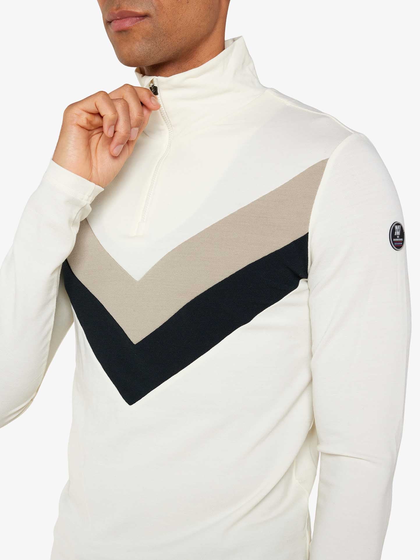 A person wearing a white Voss Chevron Zipup Men by We Norwegians with beige and black chevron patterns across the chest, made from superfine merino wool, is adjusting the partially open zipper. The zip-up features a circular patch on the left upper sleeve, adding retro vibes against a plain white background.