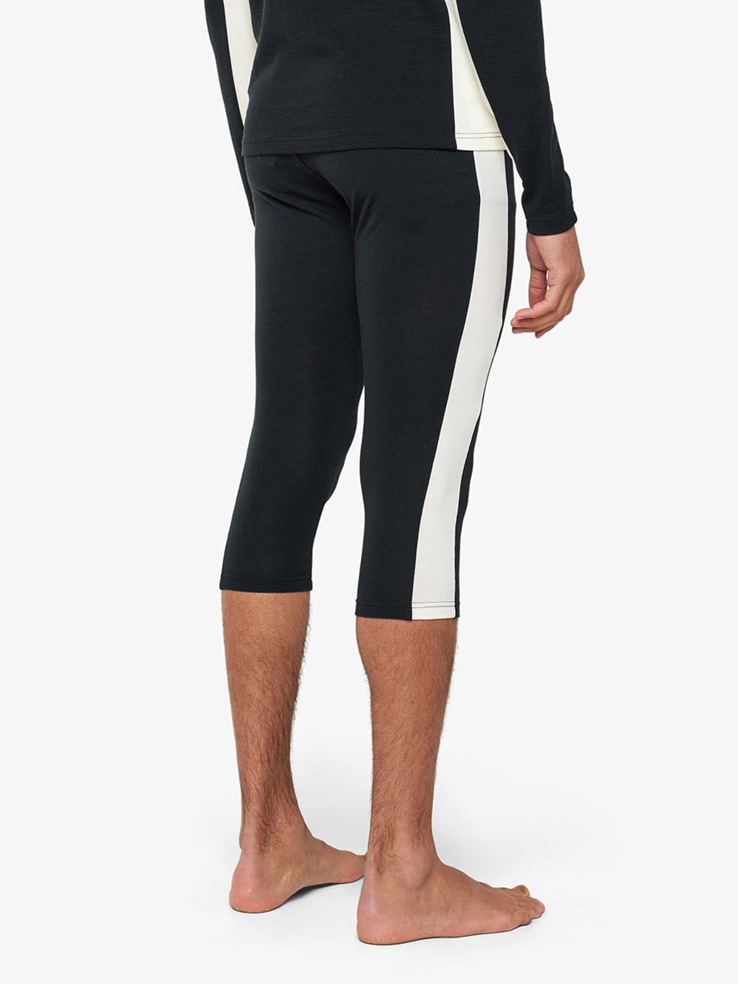 A person is standing barefoot, with their back facing the camera. They are wearing tight-fit **We Norwegians** black three-quarter-length leggings, known as **Voss Long Johns Bottoms Men**, which feature white vertical stripes on the sides and a black long-sleeve shirt with white cuffs. The background is plain white.