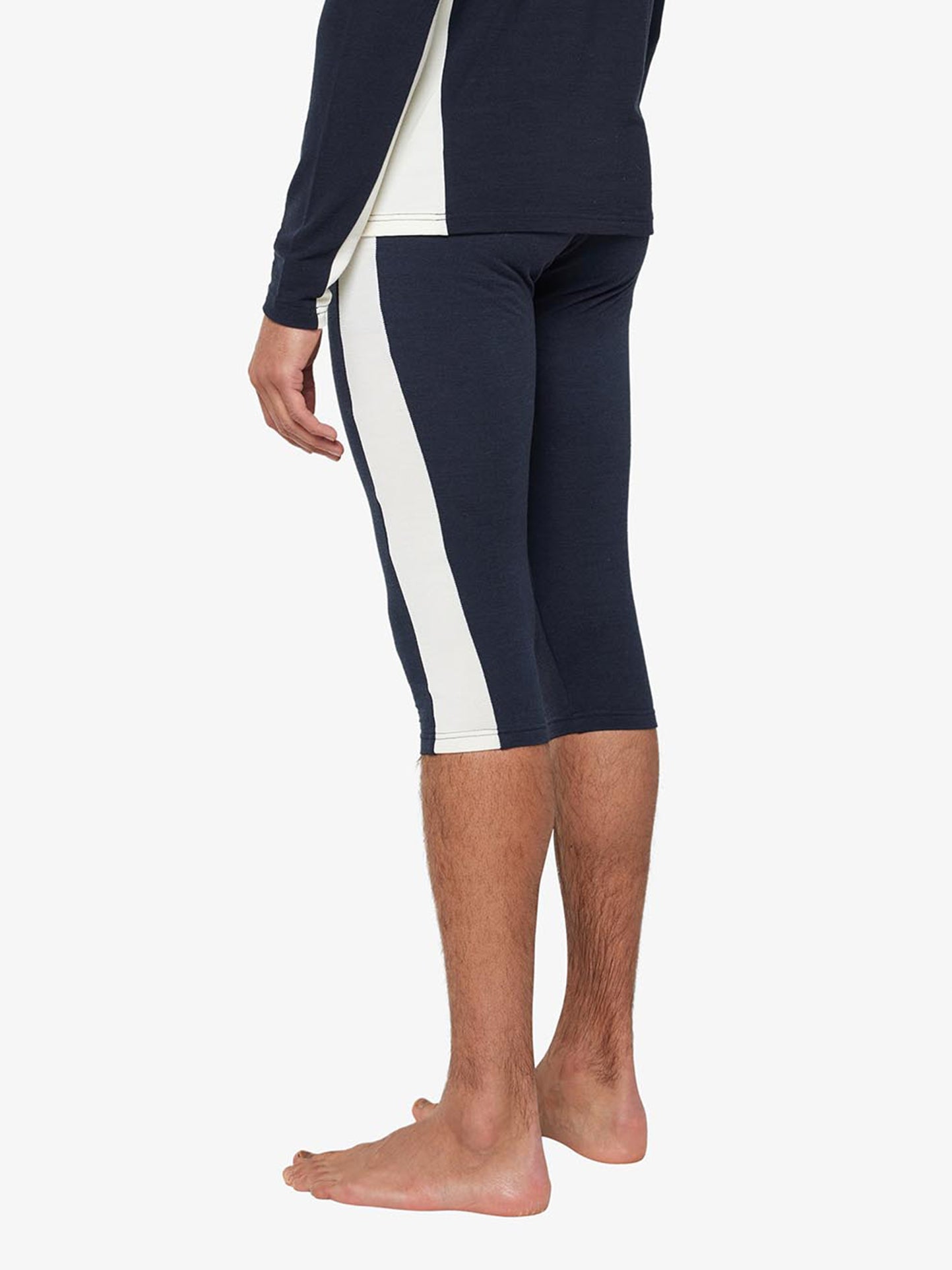 A person standing barefoot, wearing the Voss Long Johns Bottoms Men by We Norwegians, which feature navy blue athletic leggings with a wide white stripe running down the side. The individual is shown from the side, with legs bent slightly at the knee. The upper torso and arms are partially visible, dressed in a matching Merino wool base layer top.