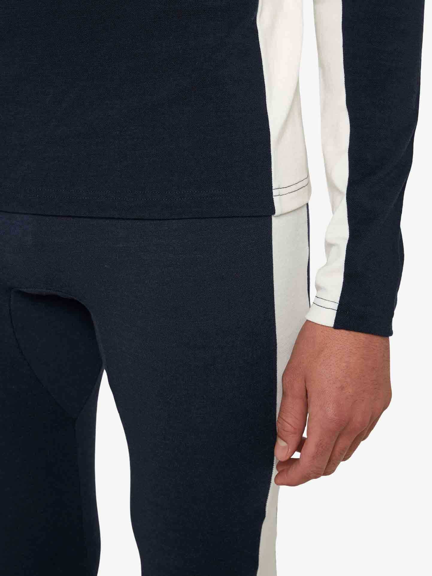 A close-up image displays the lower torso and right arm of a person wearing the Voss Long Johns Bottoms Men by We Norwegians. This fitted, two-tone outfit is crafted from merino wool and features a predominantly black design with a white stripe running down the side of both the shirt and pants. The person's relaxed hand is visible in the frame.