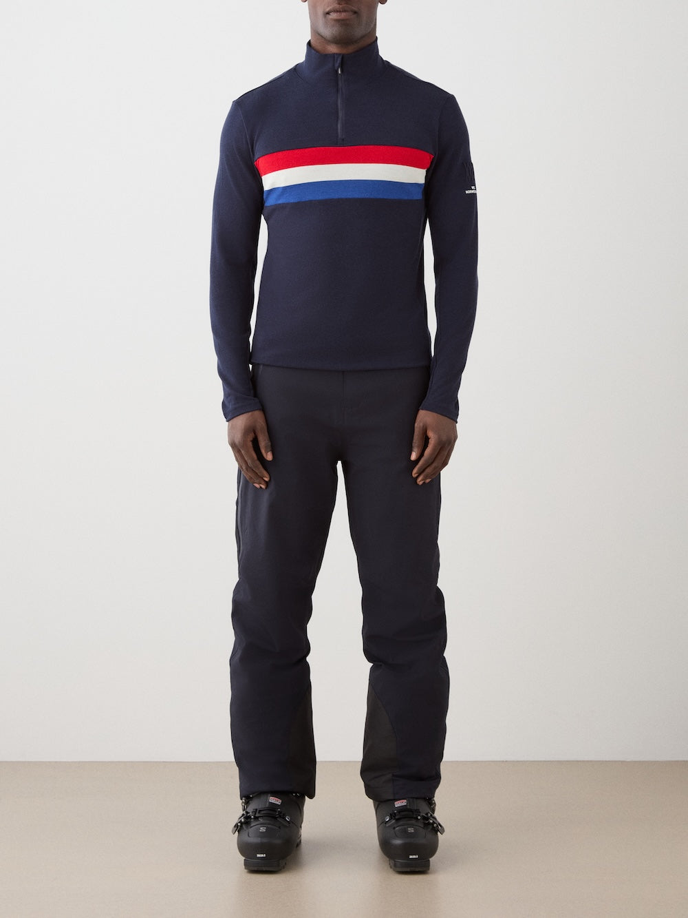 A person stands against a plain background wearing the We Norwegians Ski 1/4-Zip Men, featuring red, white, and blue chest stripes, paired with dark pants and black ski boots.